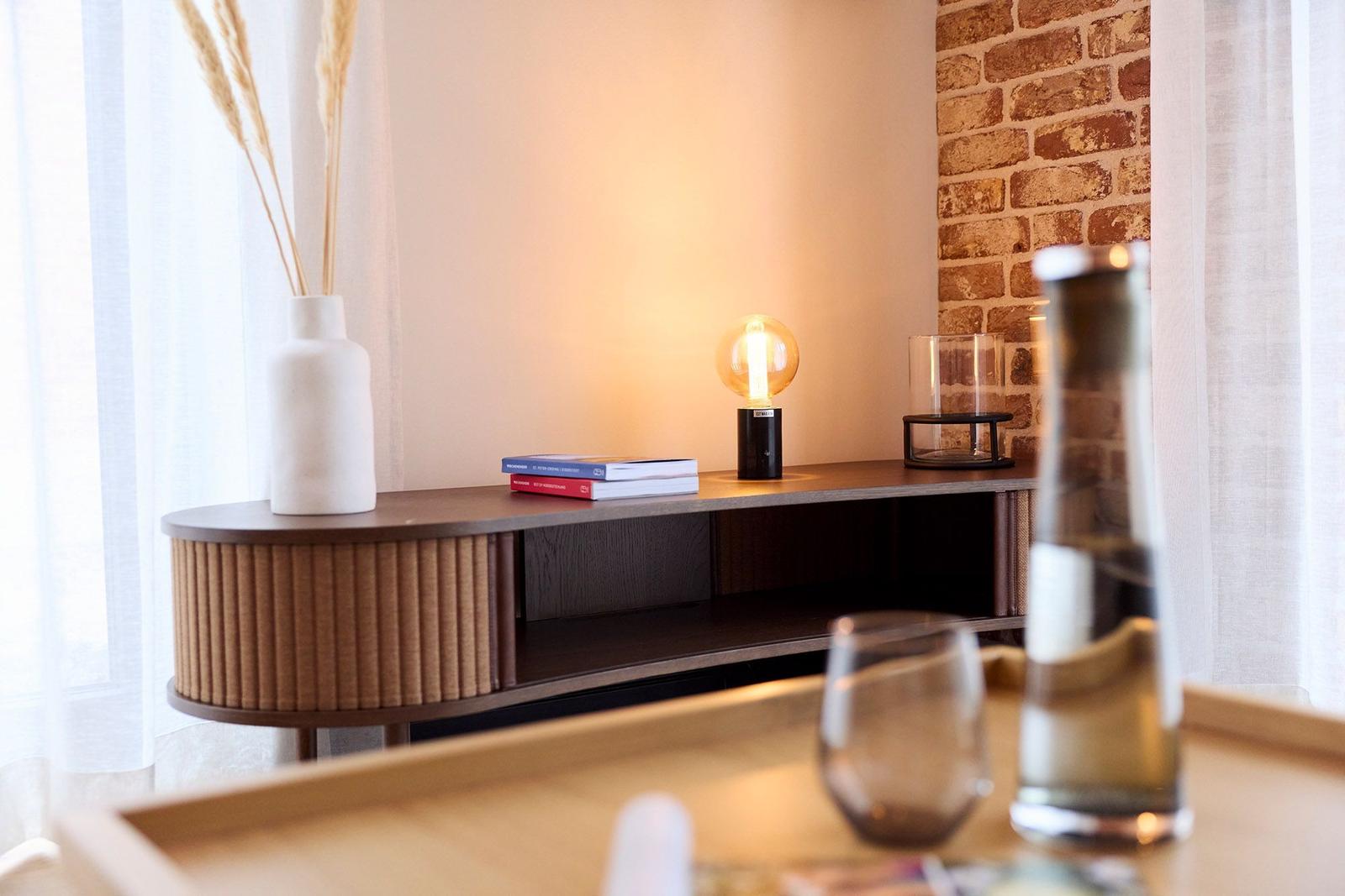 Ein Sideboard mit Lampe, Büchern und Vasen vor einer Ziegelwand.
