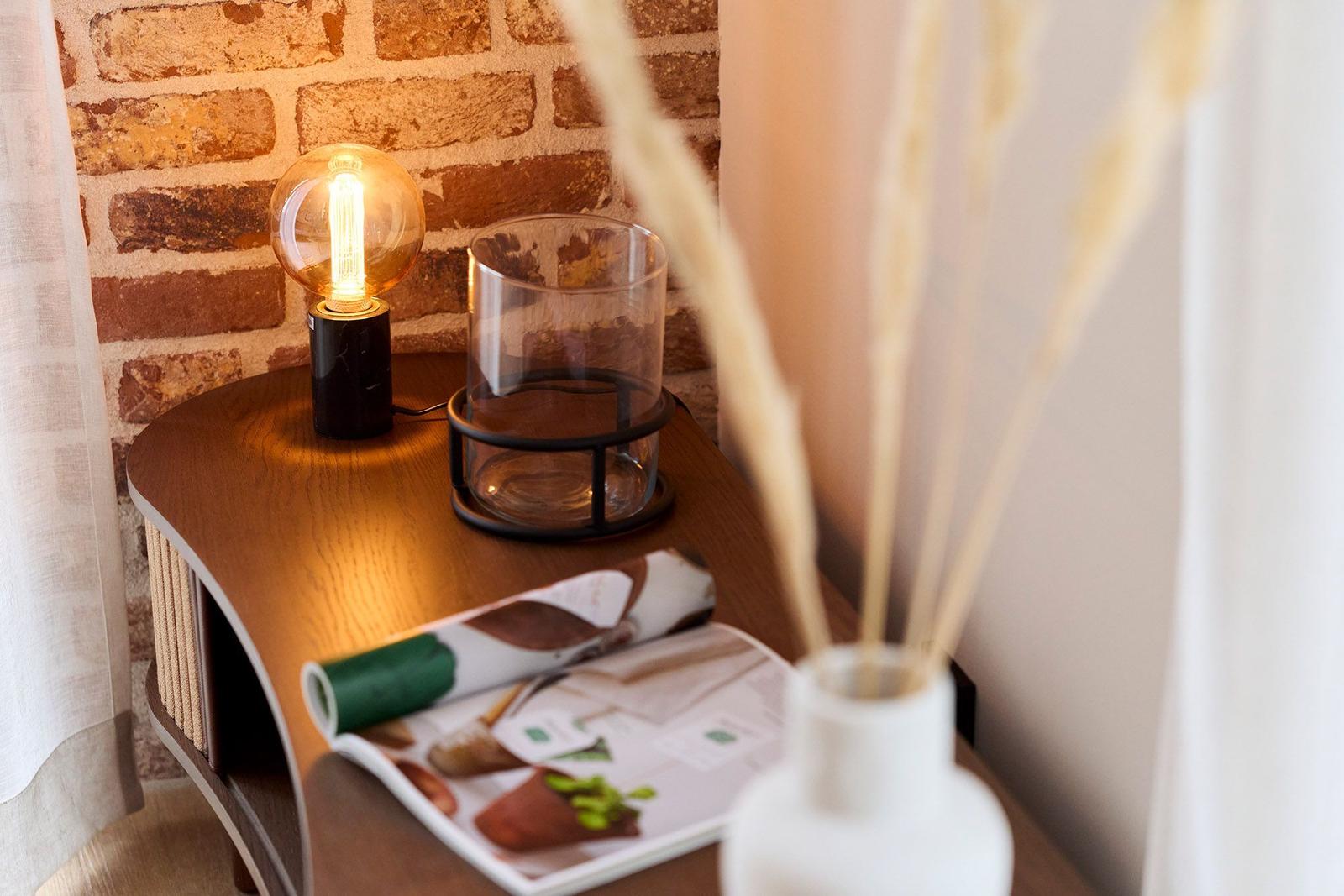 Holztisch mit Lampe, Glas und Zeitschrift vor Backsteinwand.