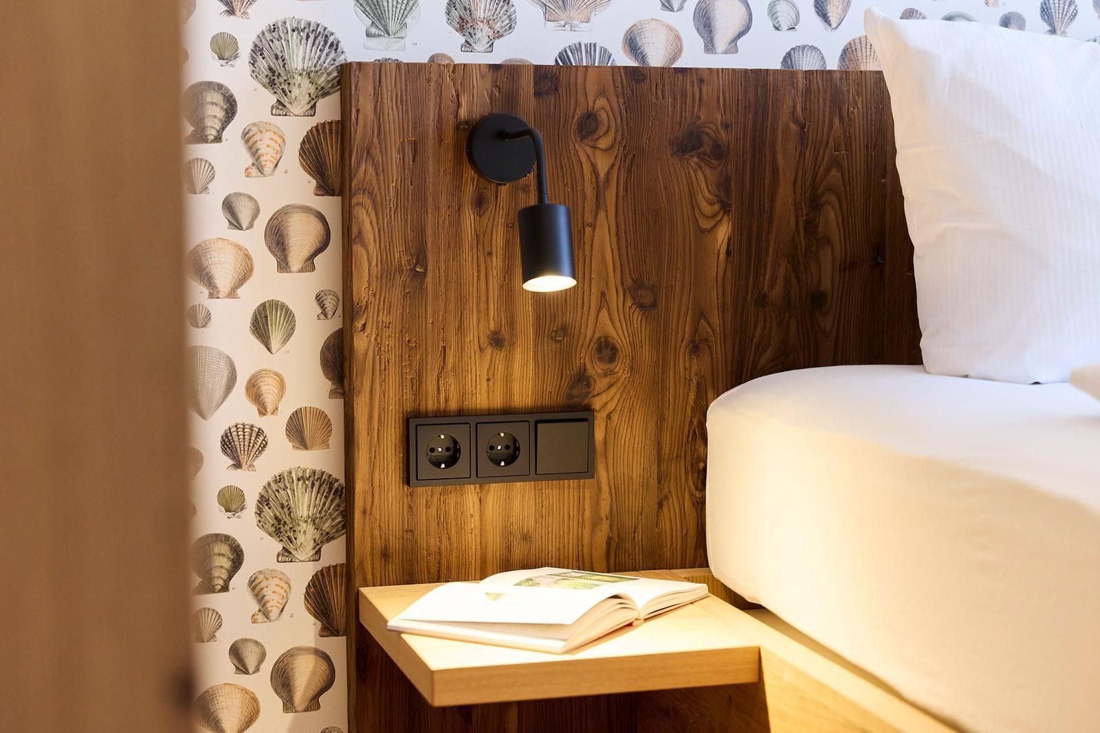 Bettkopfteil aus Holz mit Steckdose und Wandlampe. Tapete mit Muschelmotiven. Nachttisch mit Buch.