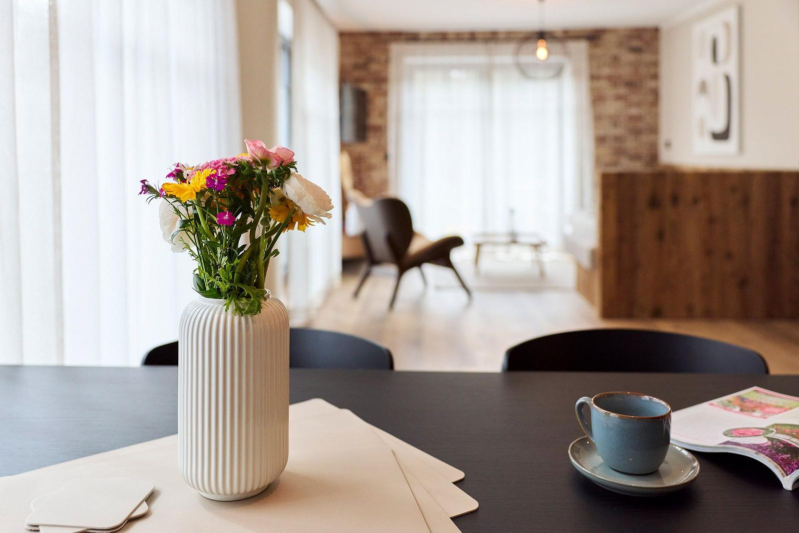 Tisch mit Blumen, Tasse und Zeitschrift im Essbereich. Im Hintergrund Sitzgelegenheit und Holzwand.