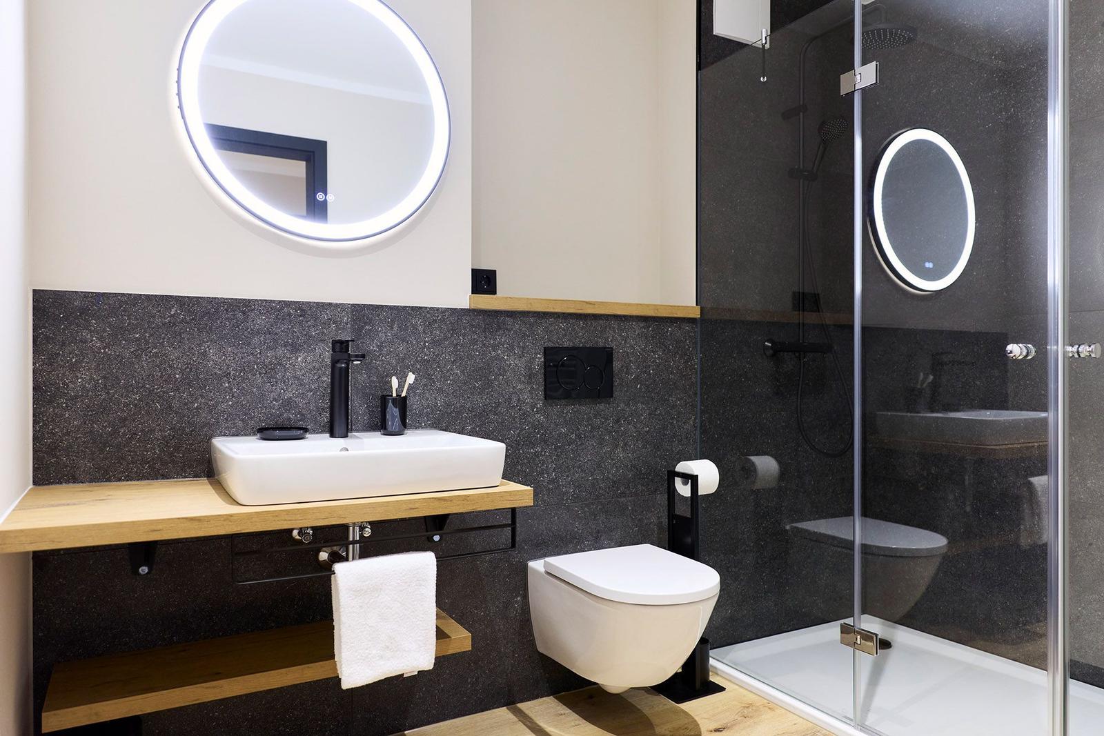 Badezimmer mit Dusche, Waschbecken und Toilette. Schwarze Wand und Holzregale.
