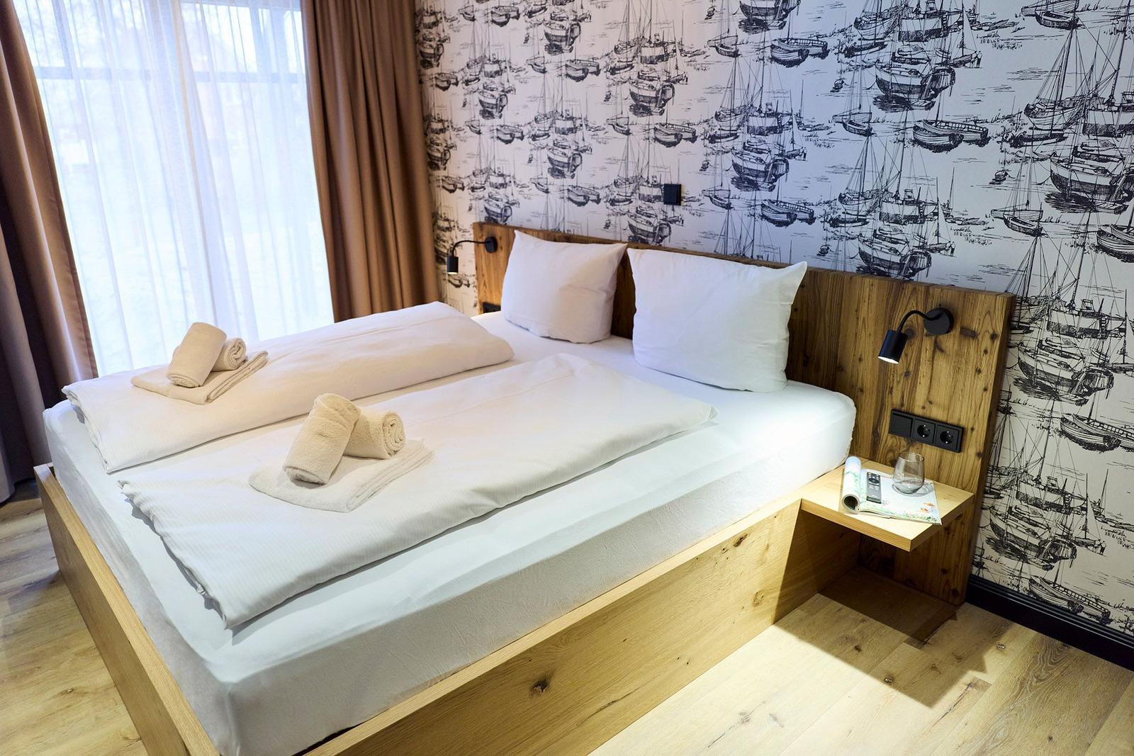 Ein Bett mit weißen Laken und Handtüchern, Holzgestell, Wand mit Booten-Motiv und Nachttisch.