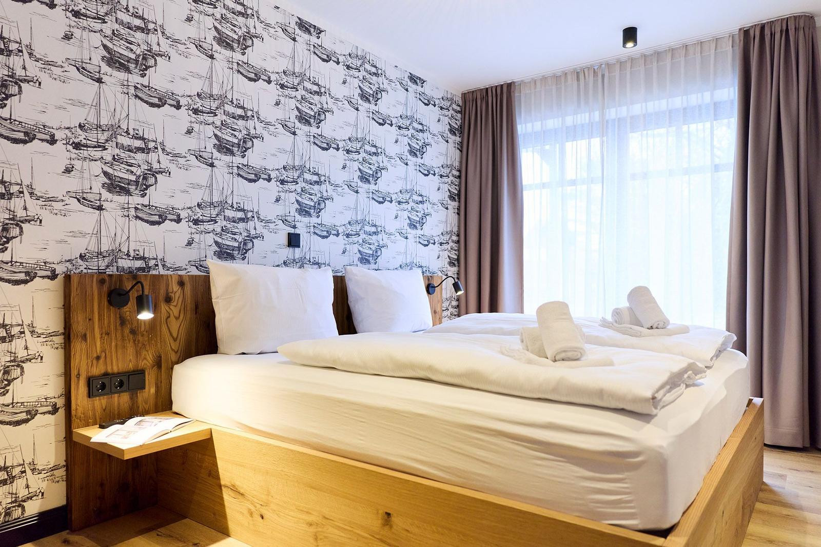 Schlafzimmer mit Holzbett, Segelyacht-Wandtapete und großem Fenster mit Vorhängen.