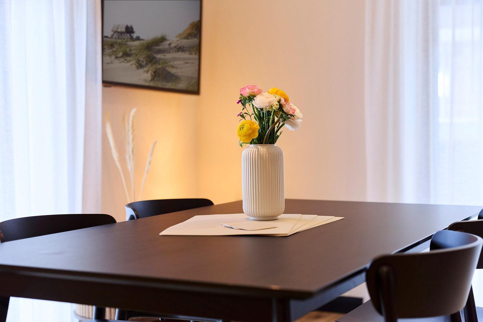 Ein Tisch mit Stühlen und einer Vase mit Blumen. An der Wand hängt ein Bild.