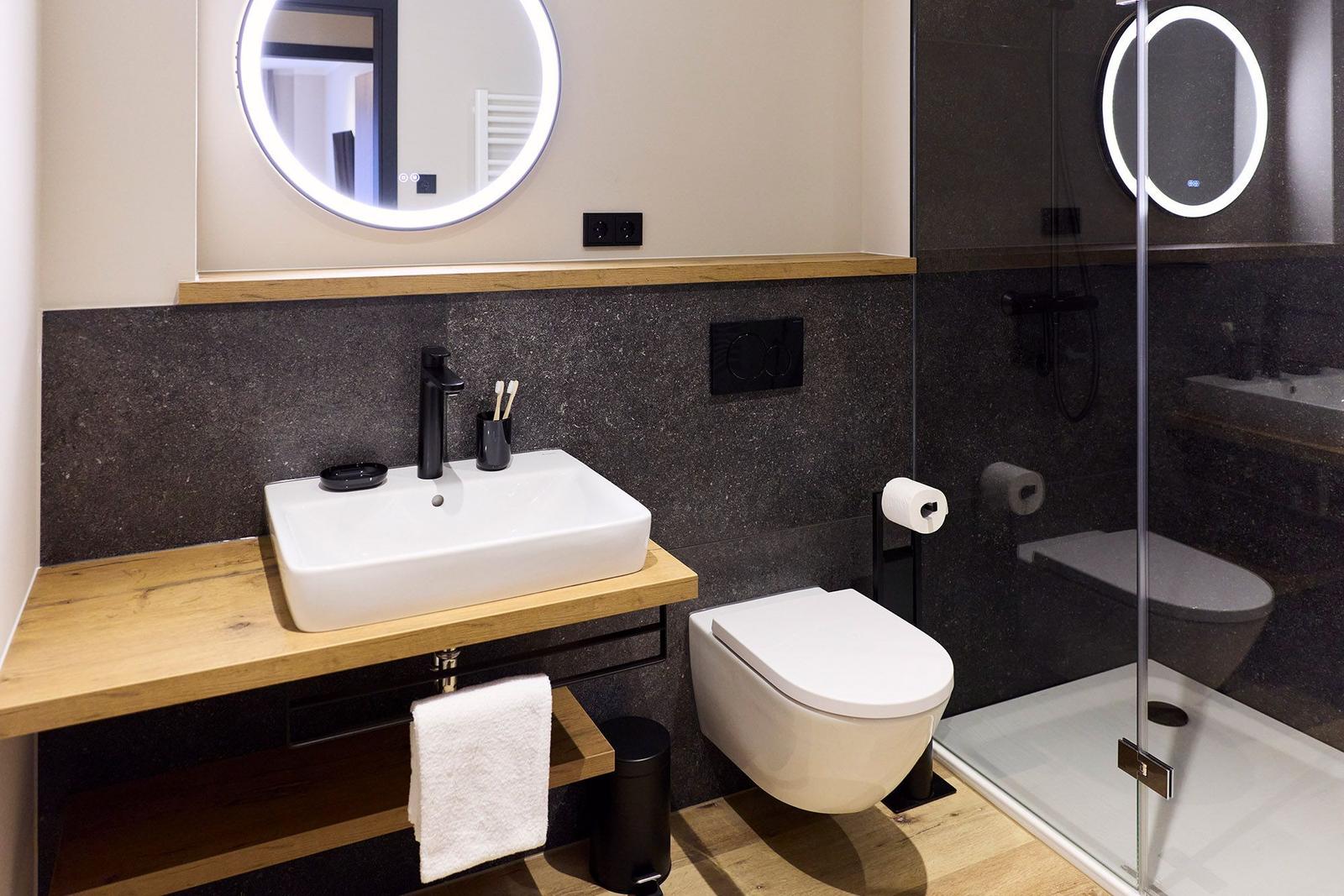 Bad mit Waschbecken, Toilette und Dusche. Runder beleuchteter Spiegel über dem Waschbecken.
