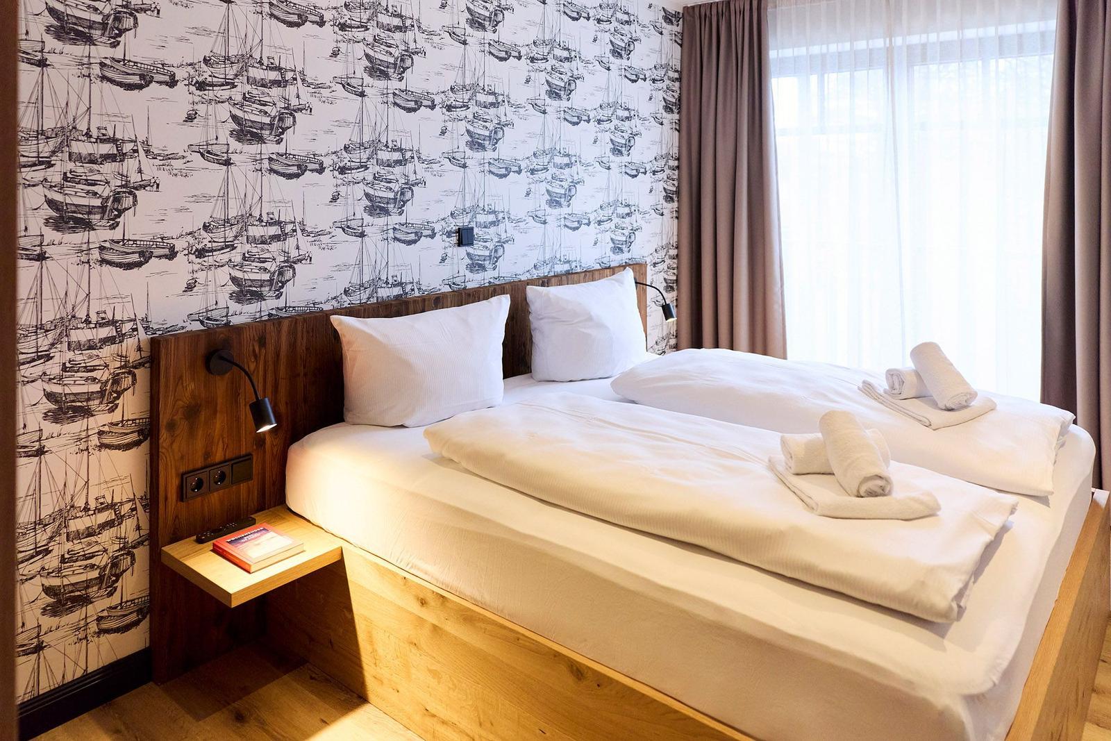 Schlafzimmer mit Bett, Wandtapete mit Schiffsmotiven und Fenster mit Vorhängen.