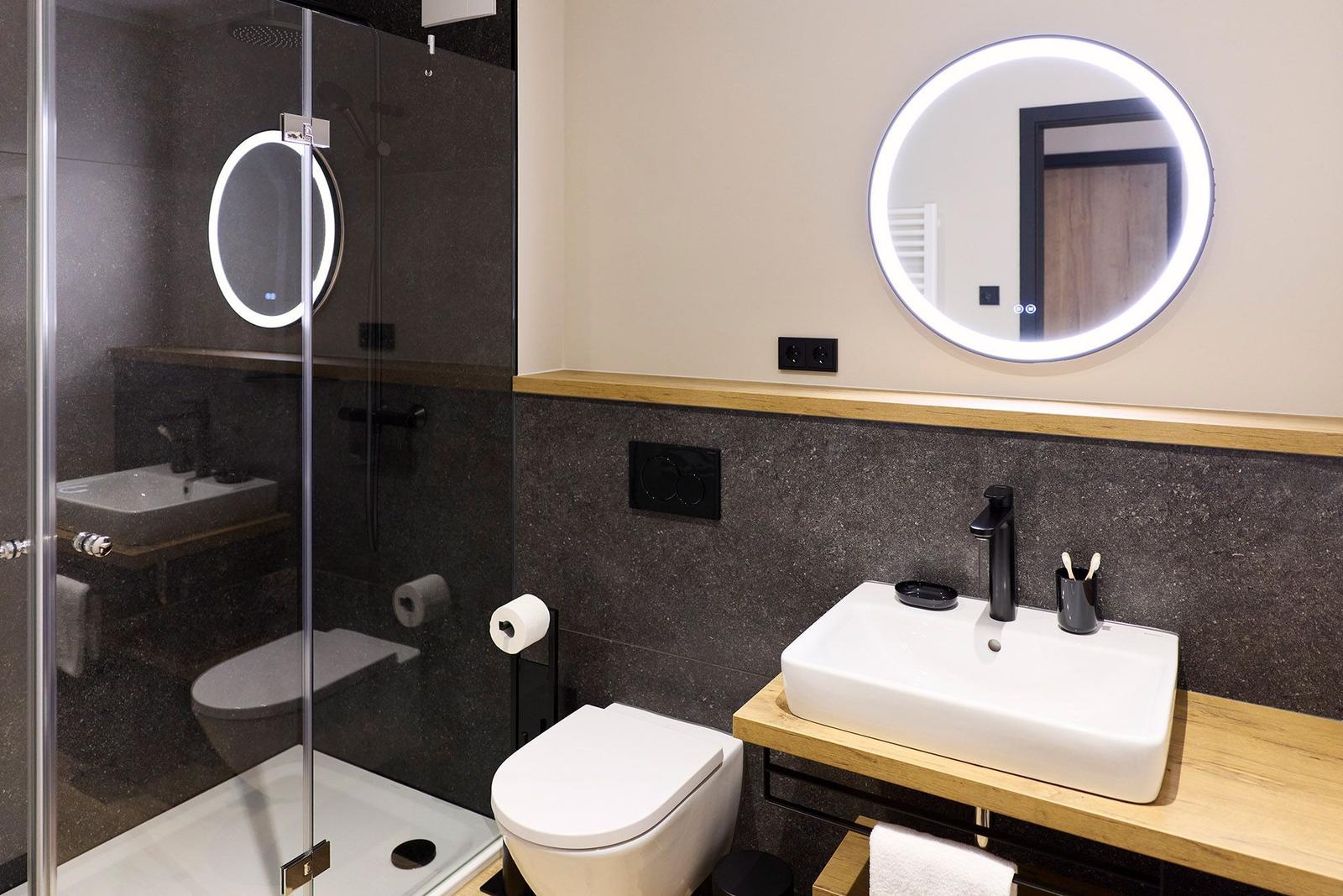 Badezimmer mit Dusche, Waschbecken und Toilette. Runde Spiegel mit Beleuchtung.