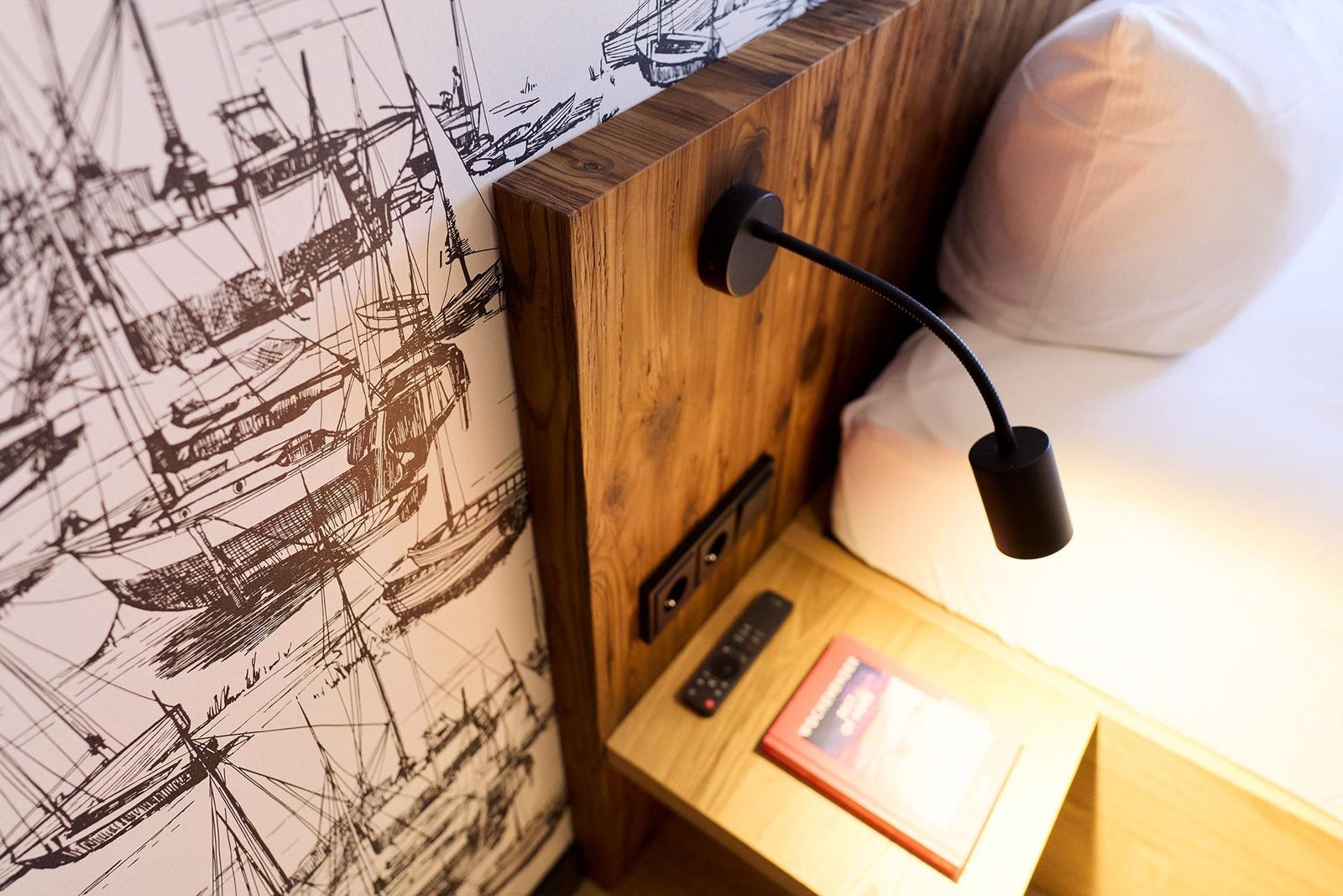 Schlafzimmer mit Holzgestell, Schiffsmotivwand und Nachttisch mit Fernbedienung und Buch.