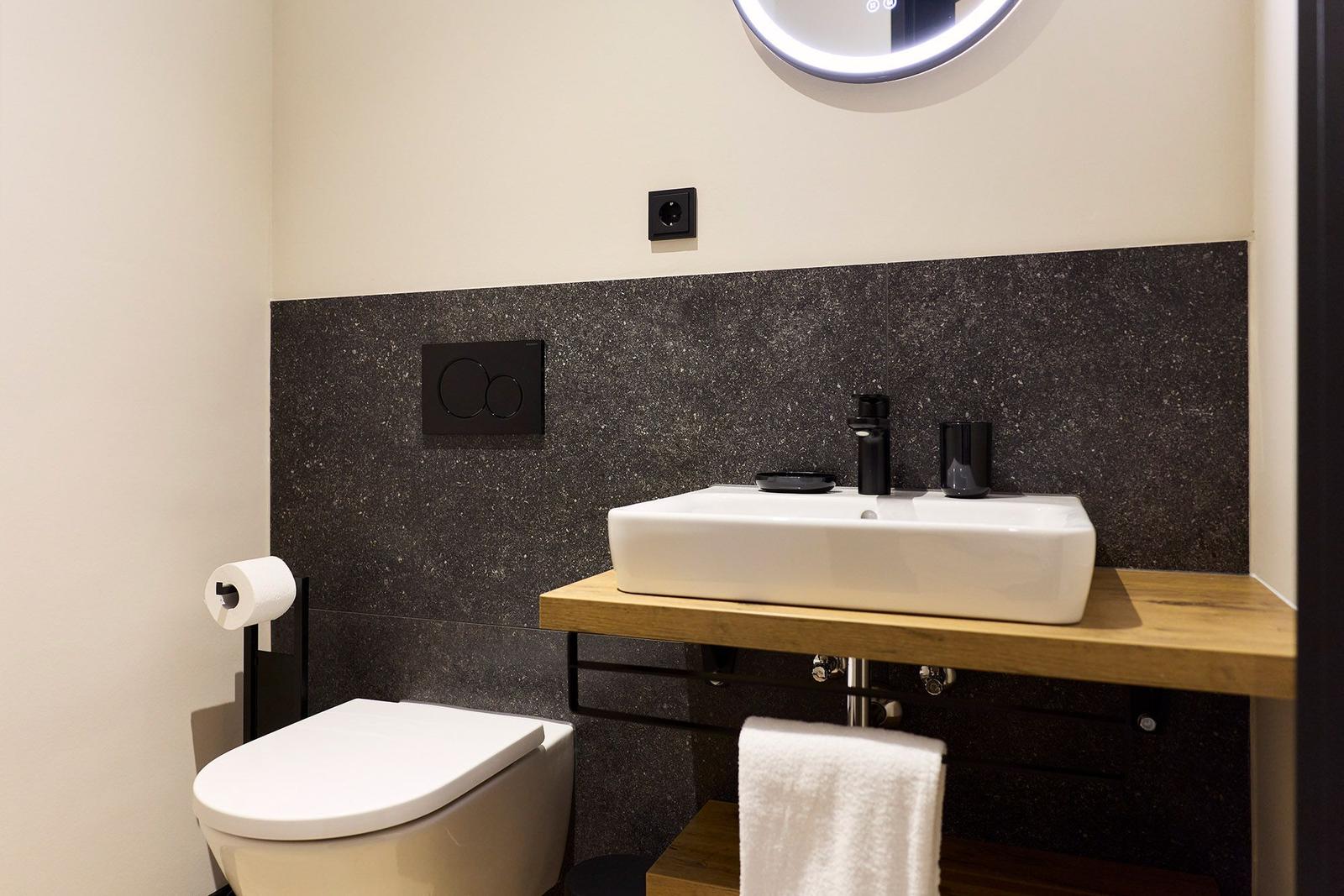 Badezimmer mit Toilette, Waschbecken und Spiegel. Schwarze Wandverkleidung und Holzunterschrank.