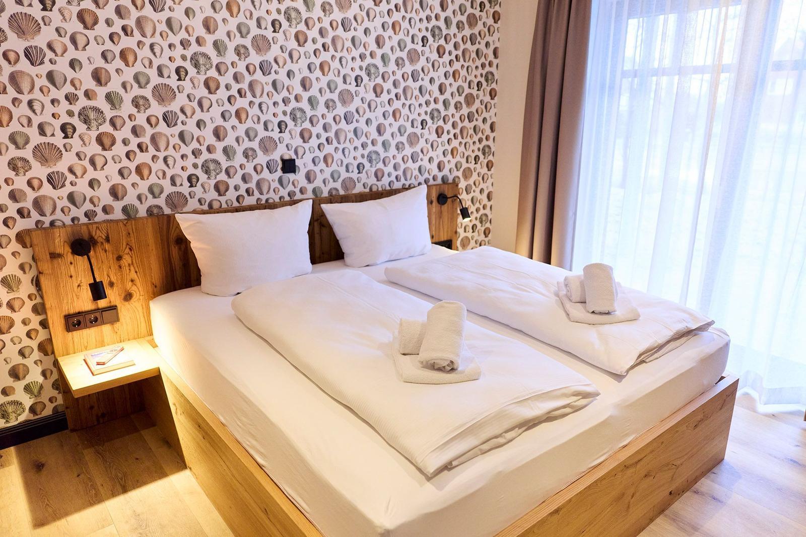 Schlafzimmer mit Doppelbett, Schalentapete und Holzgestell. Fenster mit Vorhängen.