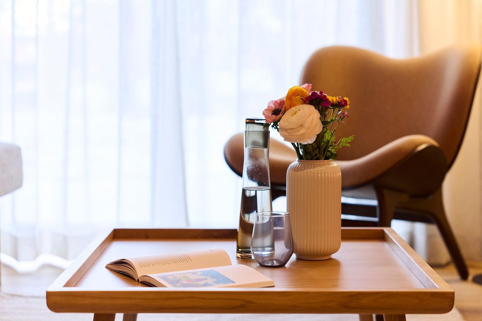 Ein Tisch mit Blumen, Wasser und einem Buch vor einem Sessel.
