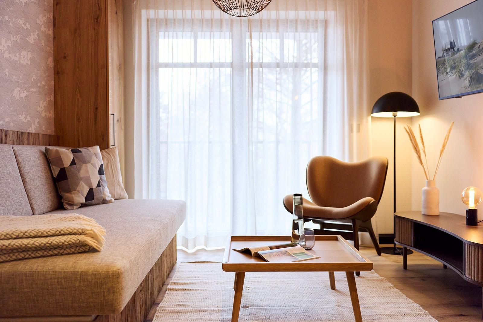 Wohnzimmer mit Sofa, Sessel, Tisch und Fernseher. Fenster mit weißen Vorhängen.