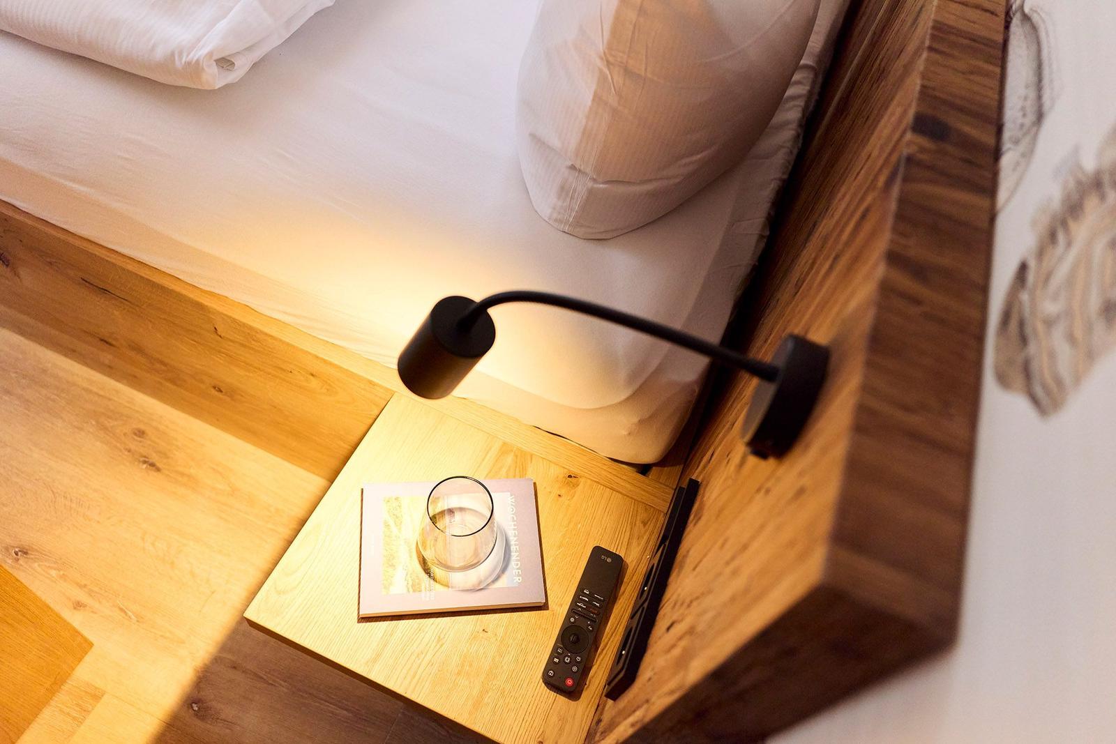 Schlafzimmer mit Holzbett, Nachttisch, Fernbedienung und Glas. Wandlampe beleuchtet die Ecke.