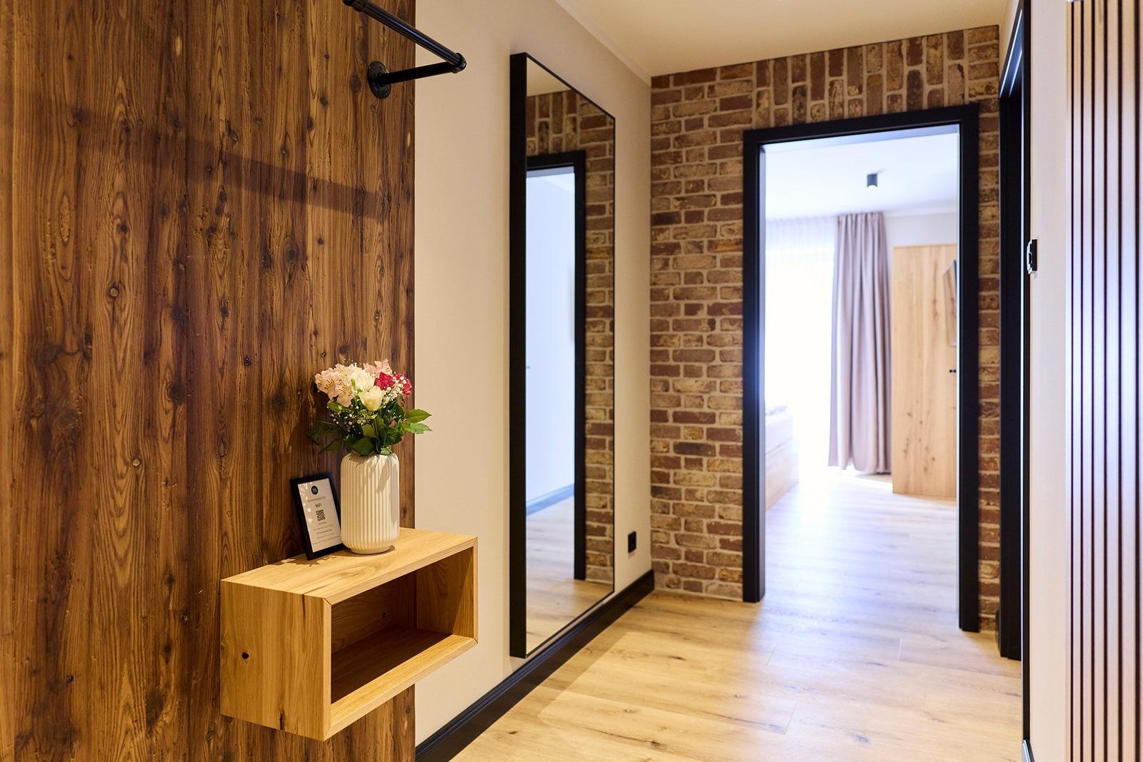 Hausgang mit Holzwand, Spiegel und Blumenarrangement. Durchgang zu einem Zimmer mit Holzboden.