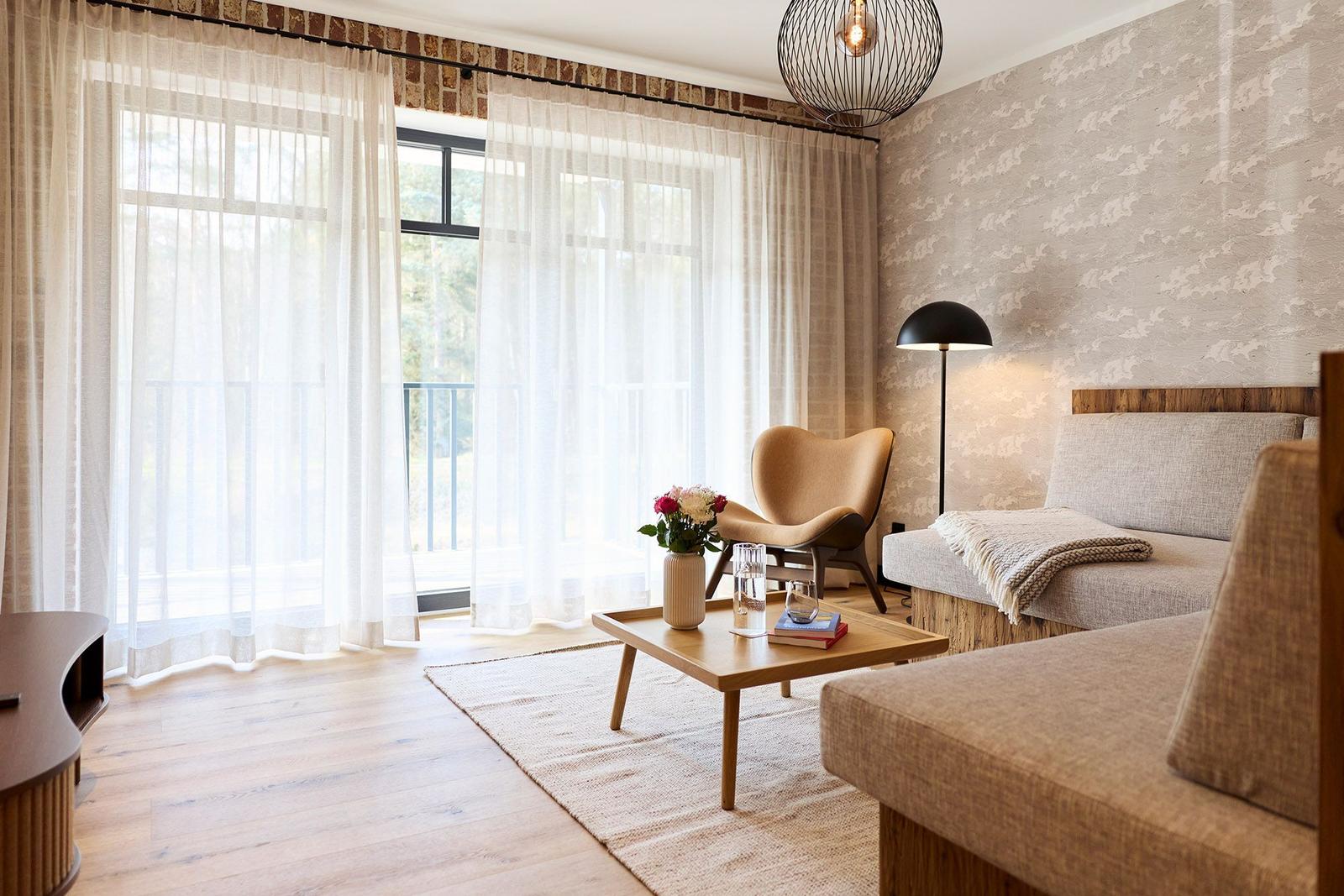 Wohnraum mit Sofa, Sessel und Tisch. Große Fenster mit Blick auf Balkon.