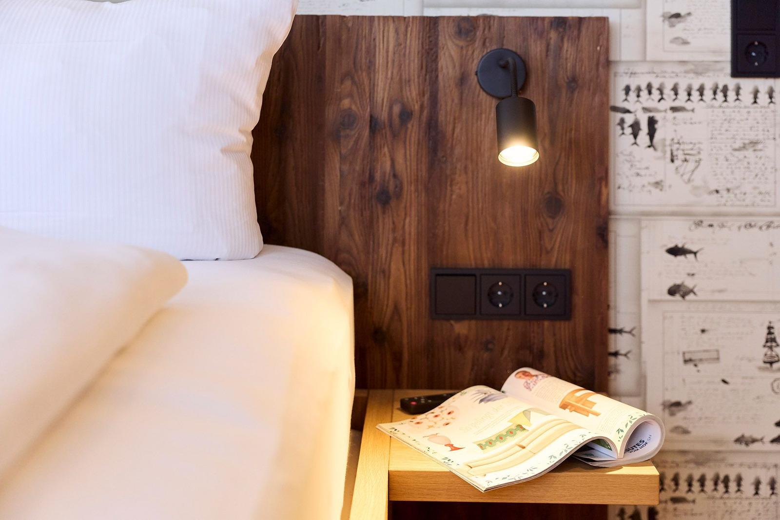 Bettkopf aus Holz mit Wandlampe und Steckdose. Auf Nachttisch liegt eine Zeitschrift.