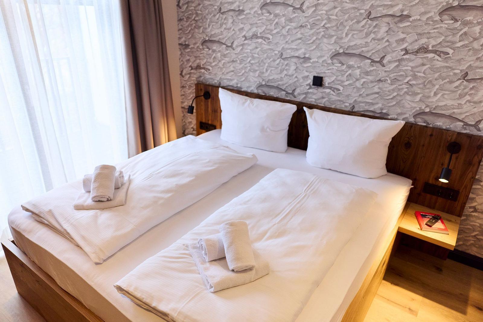 Doppelbett mit weißen Laken, zwei Kissen und Handtüchern. Wandtapete mit Fischmotiven.
