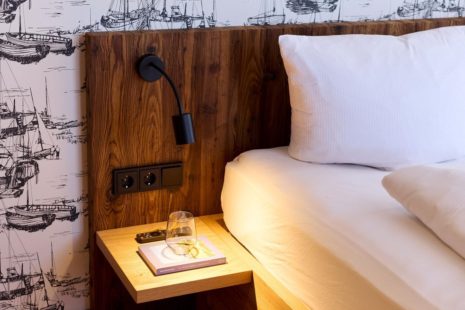 Bettkopfteil aus Holz mit Wandlampe und Steckdose. Nachttisch mit Glas und Buch. Wandtapete mit Bootsmotiven.