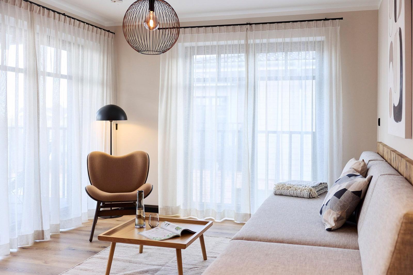 Wohnzimmer mit Sofa, Sessel, Tisch und Fenstern mit Vorhängen.