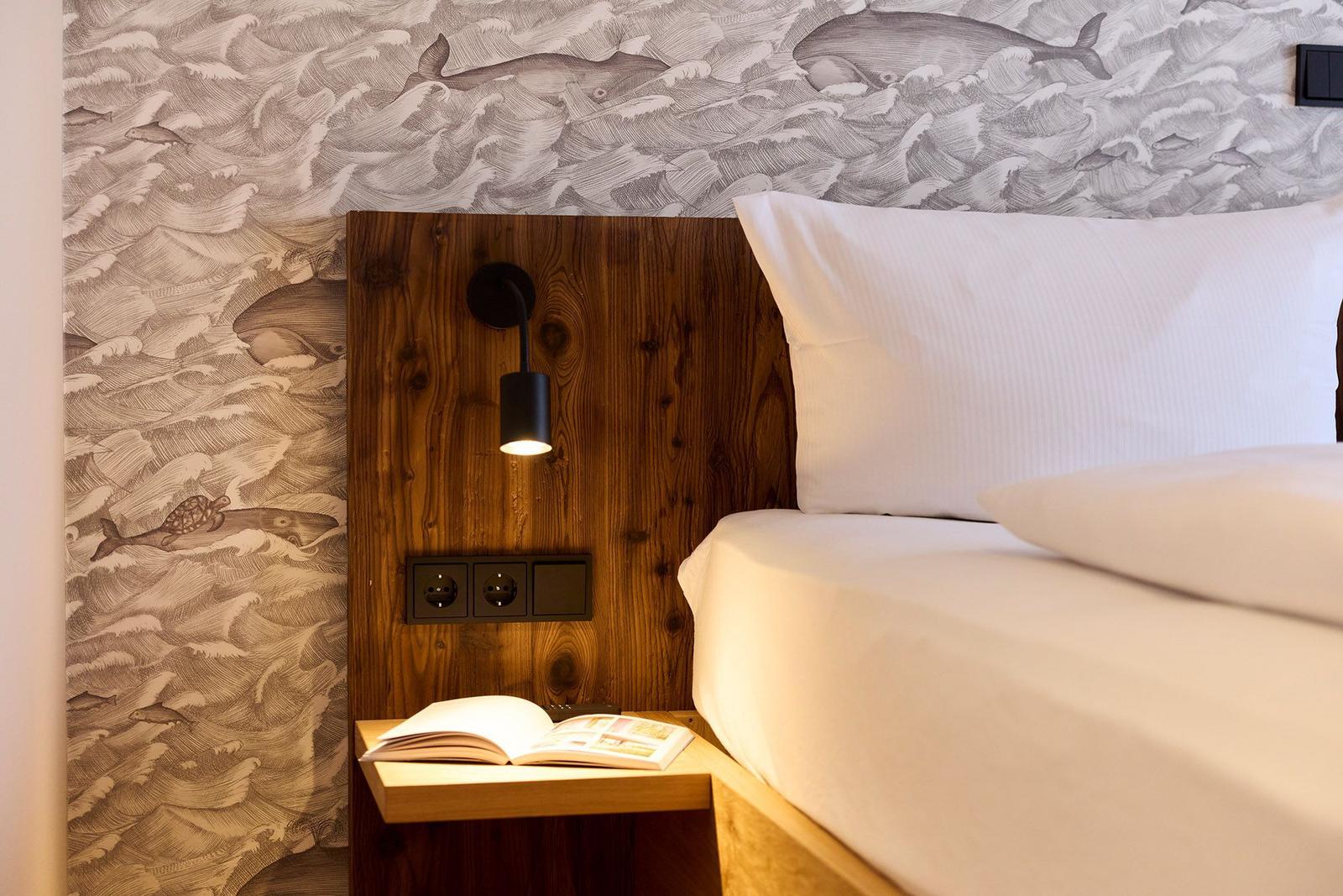 Schlafzimmer mit Holzkopfteil, Meerestier-Wandtapete und Nachttisch mit Buch.
