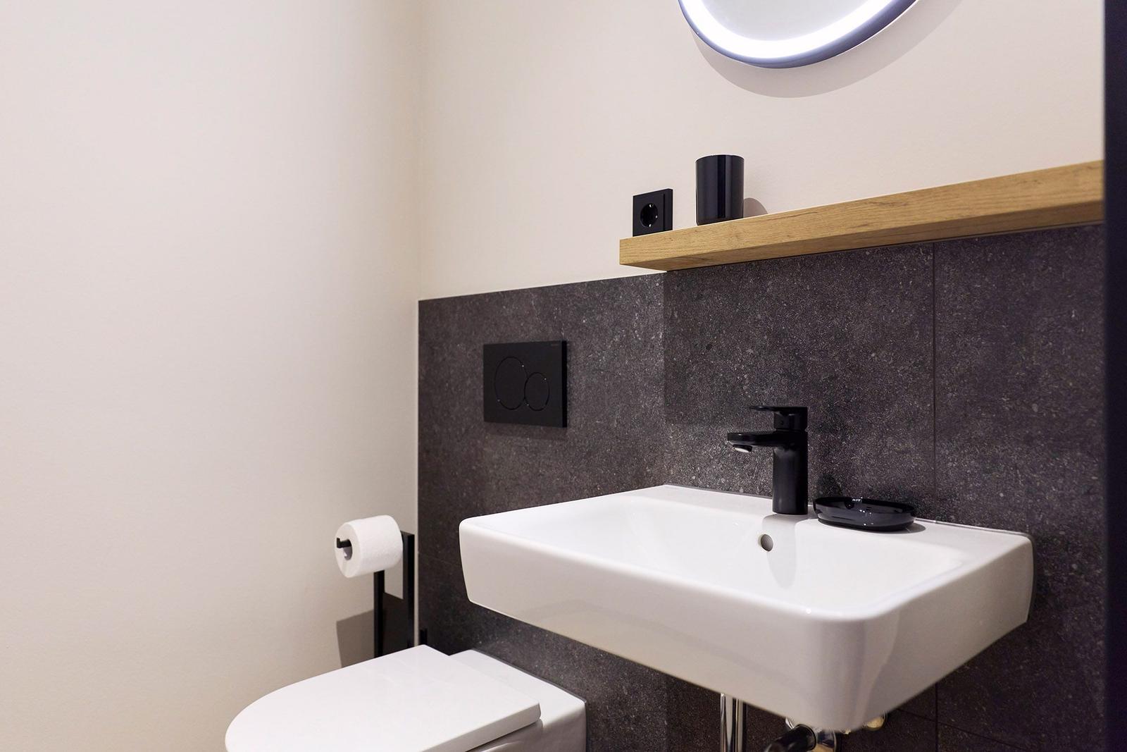 Badezimmer mit Waschbecken, Spiegel, Toilette und Holzregal an der Wand.