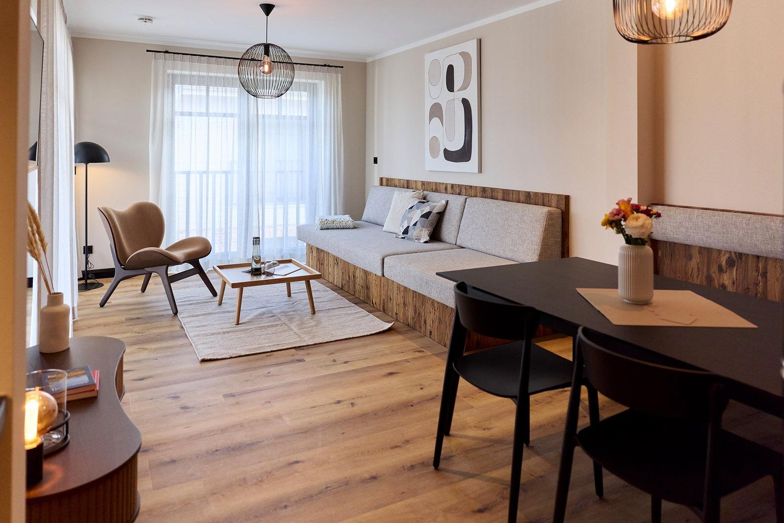 Wohn- und Essbereich mit Sofa, Tisch und Stühlen. Holzboden und Fenster mit Vorhängen.