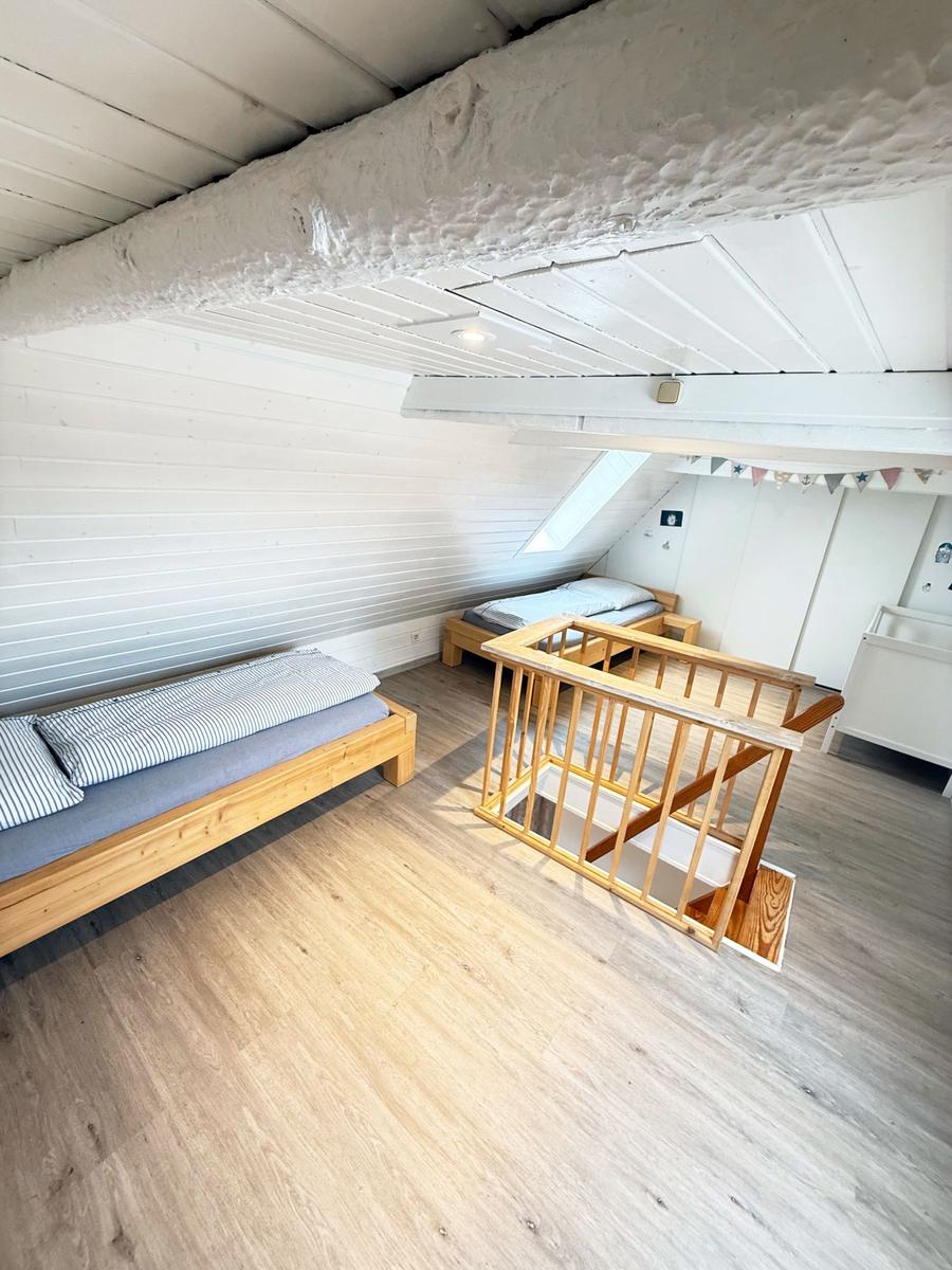 Zimmer mit zwei Betten, Holzboden und Dachschräge. Holzgeländer und Fenster sichtbar.