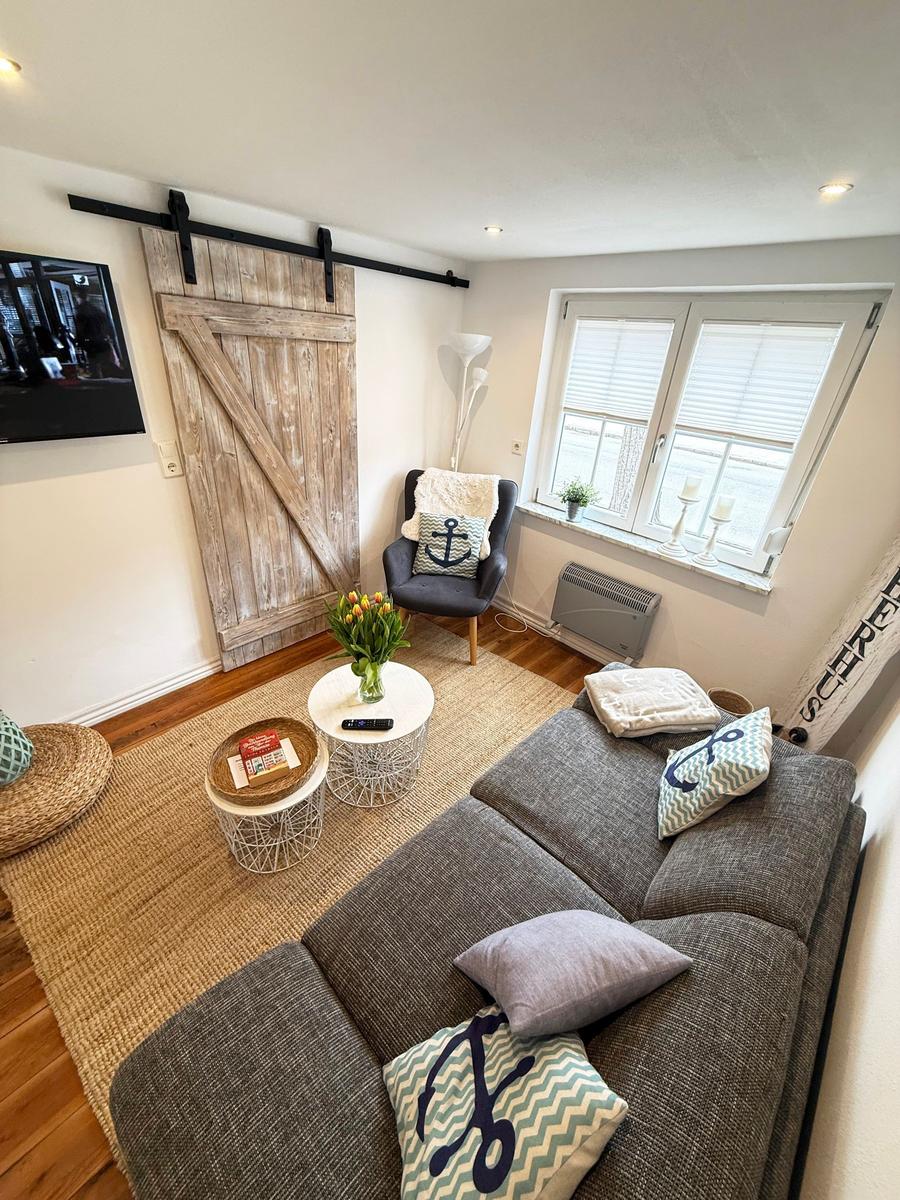 Wohnzimmer mit Sofa, Sessel, TV und Holzdecke. Fenster mit Jalousien und Blumen.