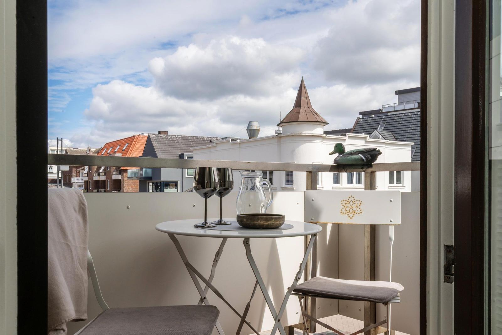 Balcony with table, chairs, and duck figurine overlooking rooftops and a tower.