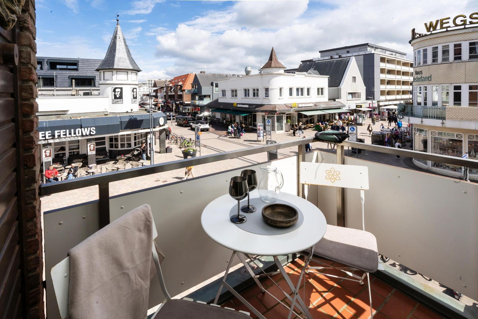 Balkon mit Tisch und Stühlen bietet Blick auf belebte Straße mit Geschäften und Passanten.