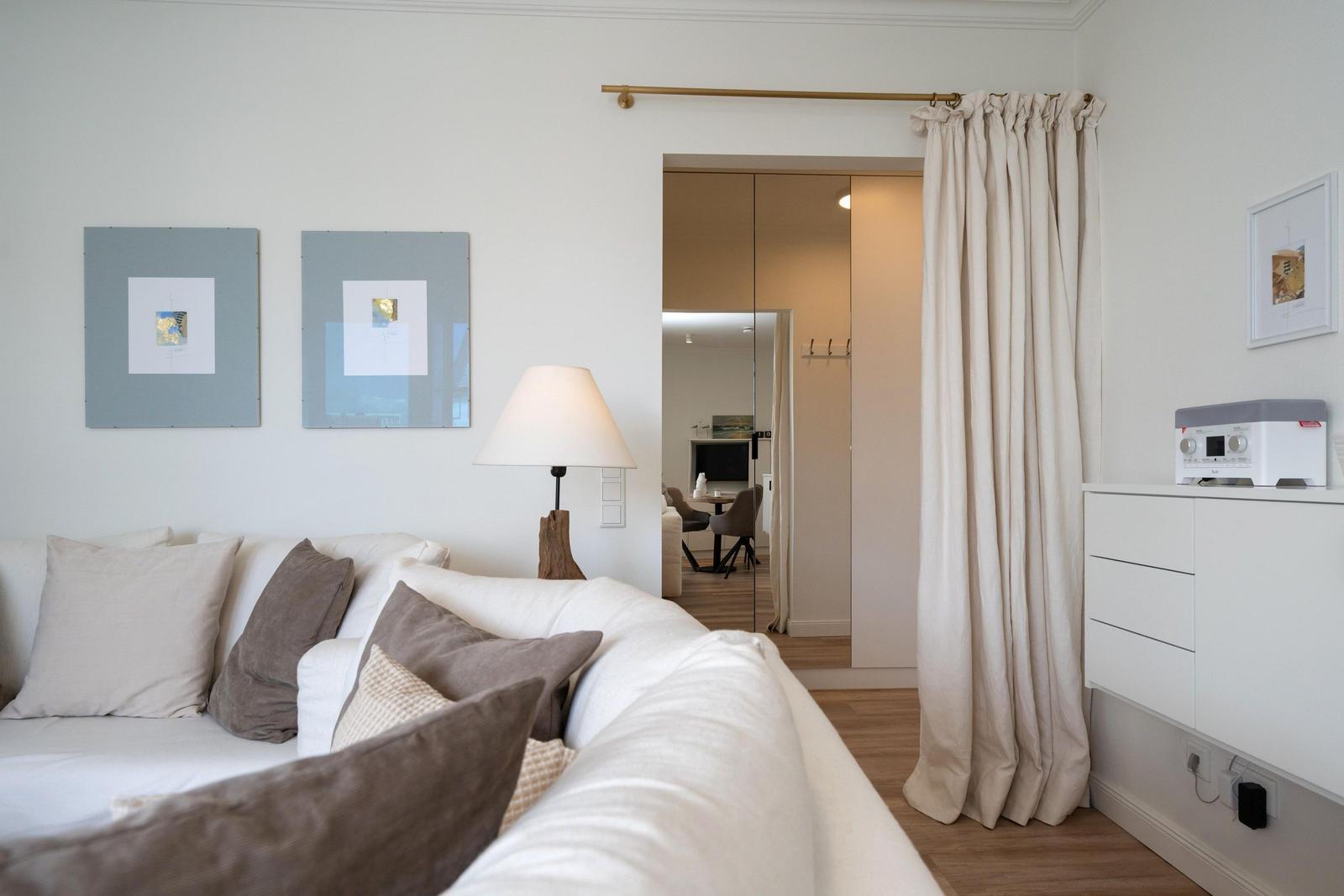 Wohnzimmer mit Sofa, Lampen und Bildern an der Wand. Durchgang zur Küche mit Vorhang.