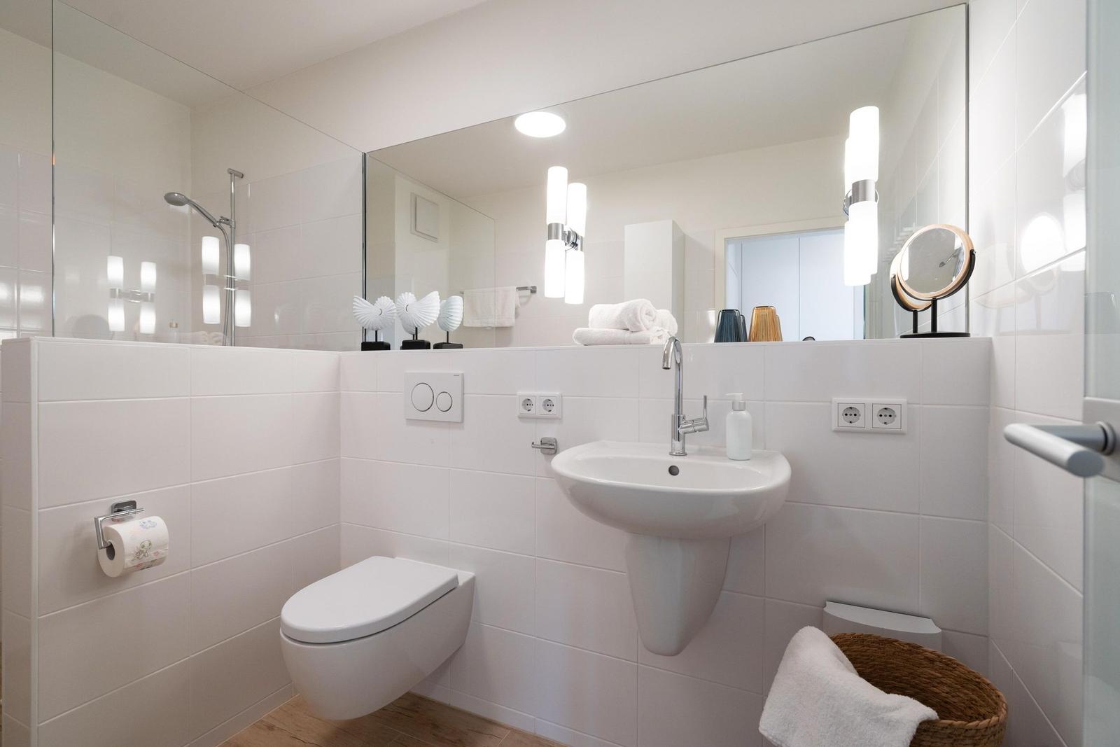 Badezimmer mit Toilette, Waschbecken und Dusche. Weiße Fliesen und Spiegel.