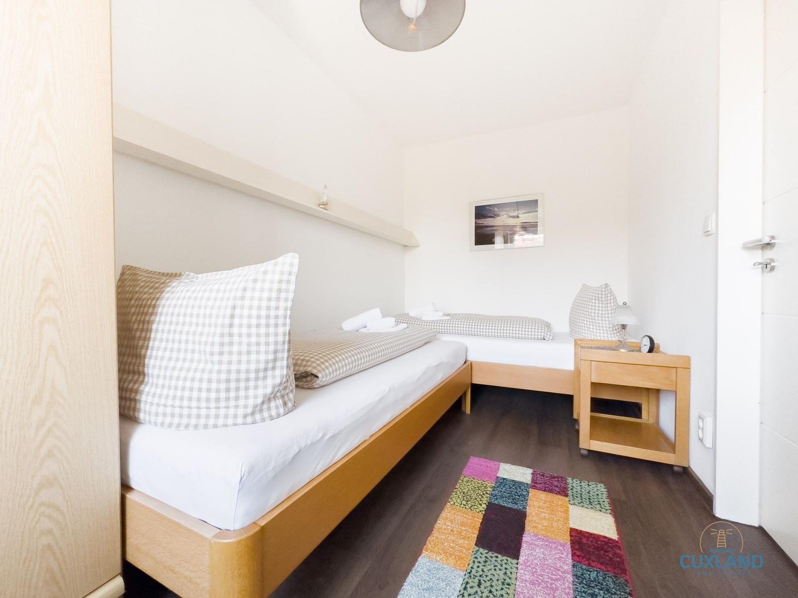 Zimmer mit zwei Betten, Holzmöbeln und buntem Teppich auf dunklem Holzfußboden.
