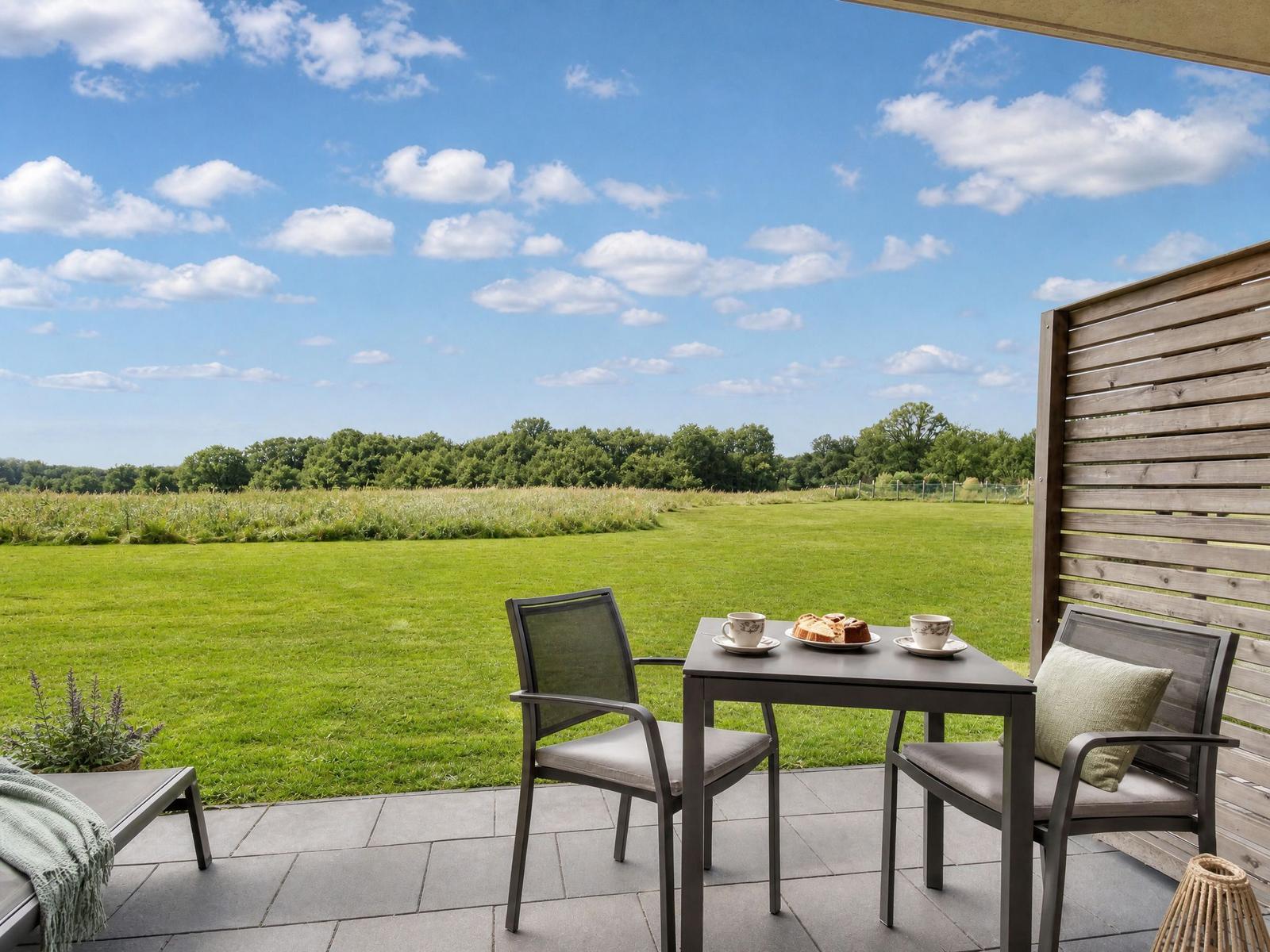 Terrasse mit Tisch, zwei Stühlen und Blick auf grünes Feld unter blauem Himmel.