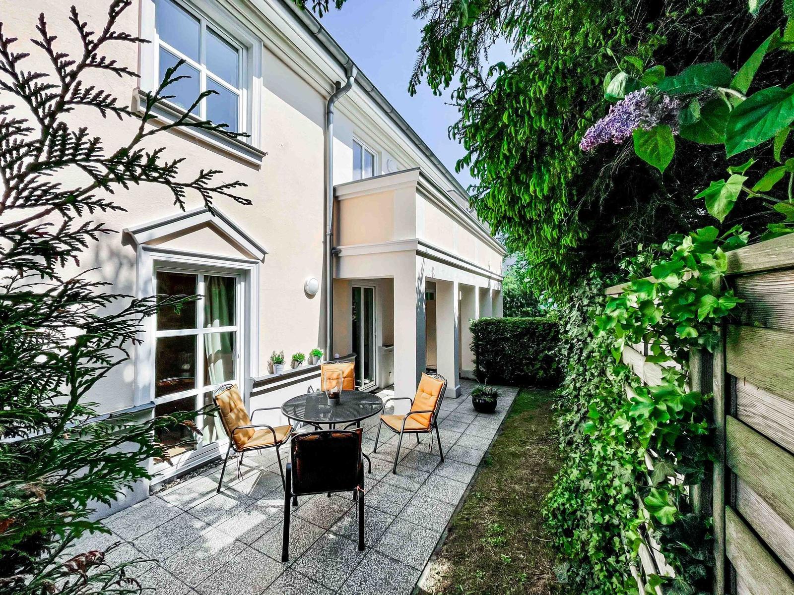 Terrasse mit Tisch und Stühlen, umgeben von grüner Vegetation und einem Holzzaun.