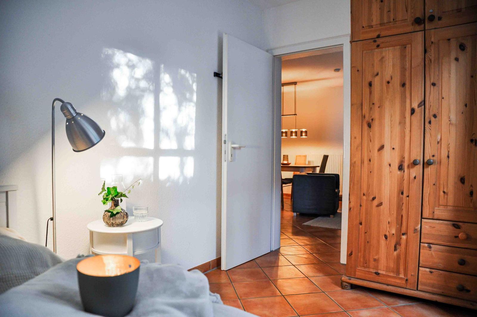 Schlafzimmer mit Blick in Wohnbereich. Holzschrank, Nachttisch und Stehlampe.