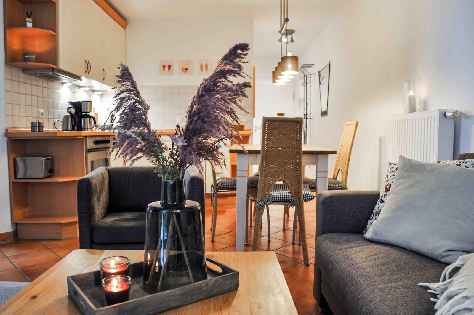 Wohnzimmer mit Esstisch, Sofa und Küchenbereich. Holztisch, Sessel und Vase mit Pflanzen.