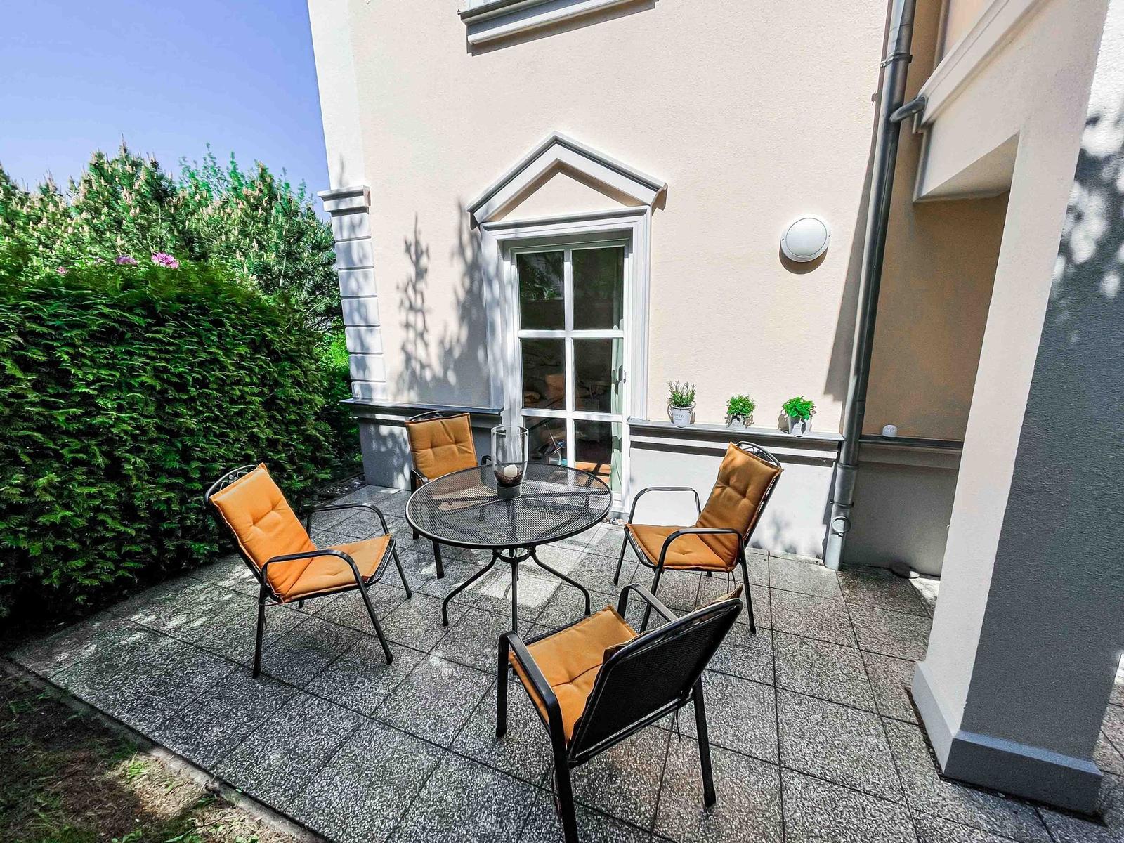 Terrasse mit Tisch und vier Stühlen, orangefarbene Sitzkissen, Pflanzen und Fenster im Hintergrund.