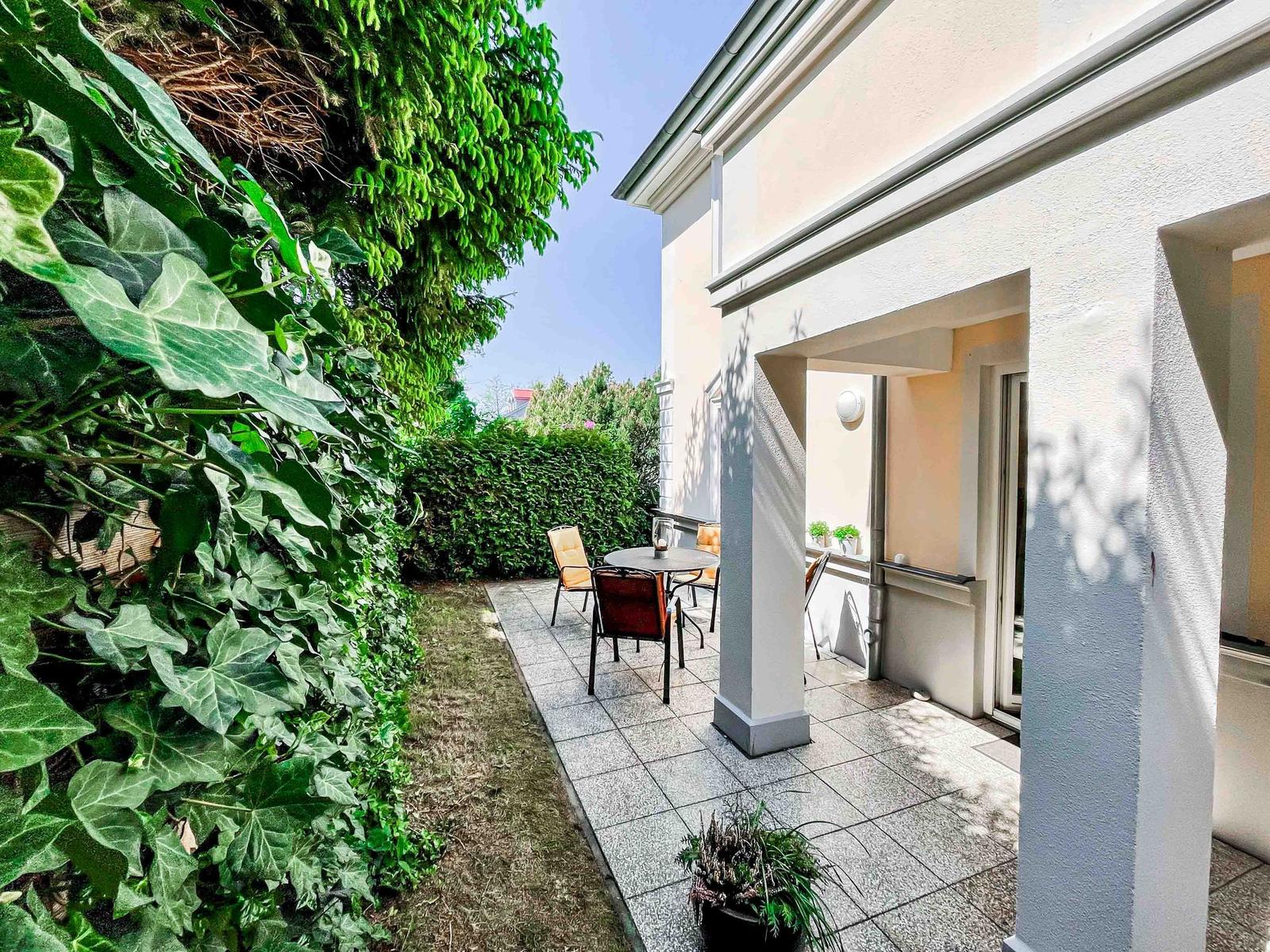 Terrasse mit Tisch und Stühlen, umgeben von grüner Vegetation und einem weißen Gebäude.