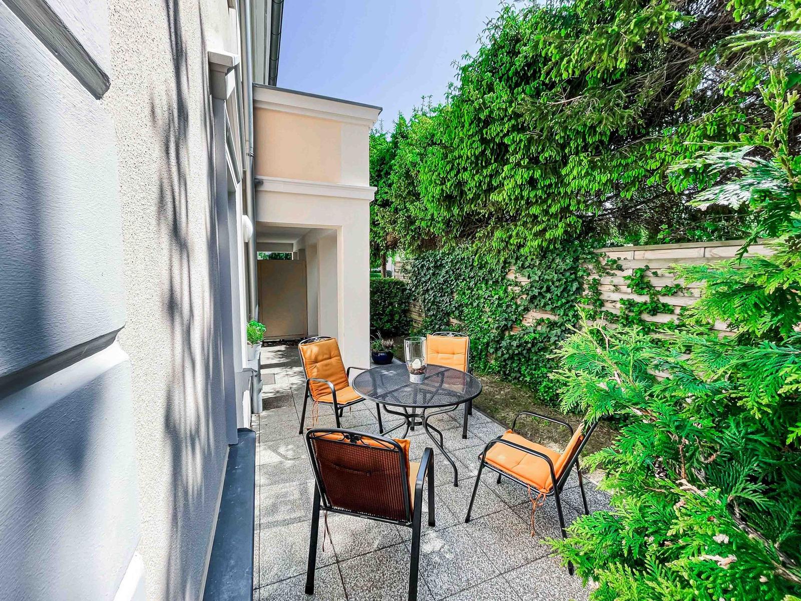 Terrasse mit Tisch und vier Stühlen, umgeben von grünen Pflanzen und einer Steinmauer.