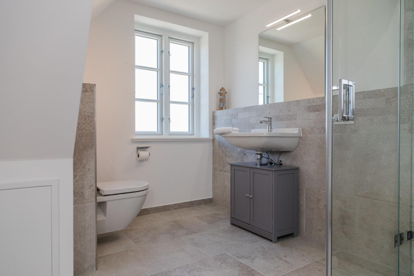 Badezimmer mit Dusche, Waschbecken und Toilette. Fenster mit weißem Rahmen.