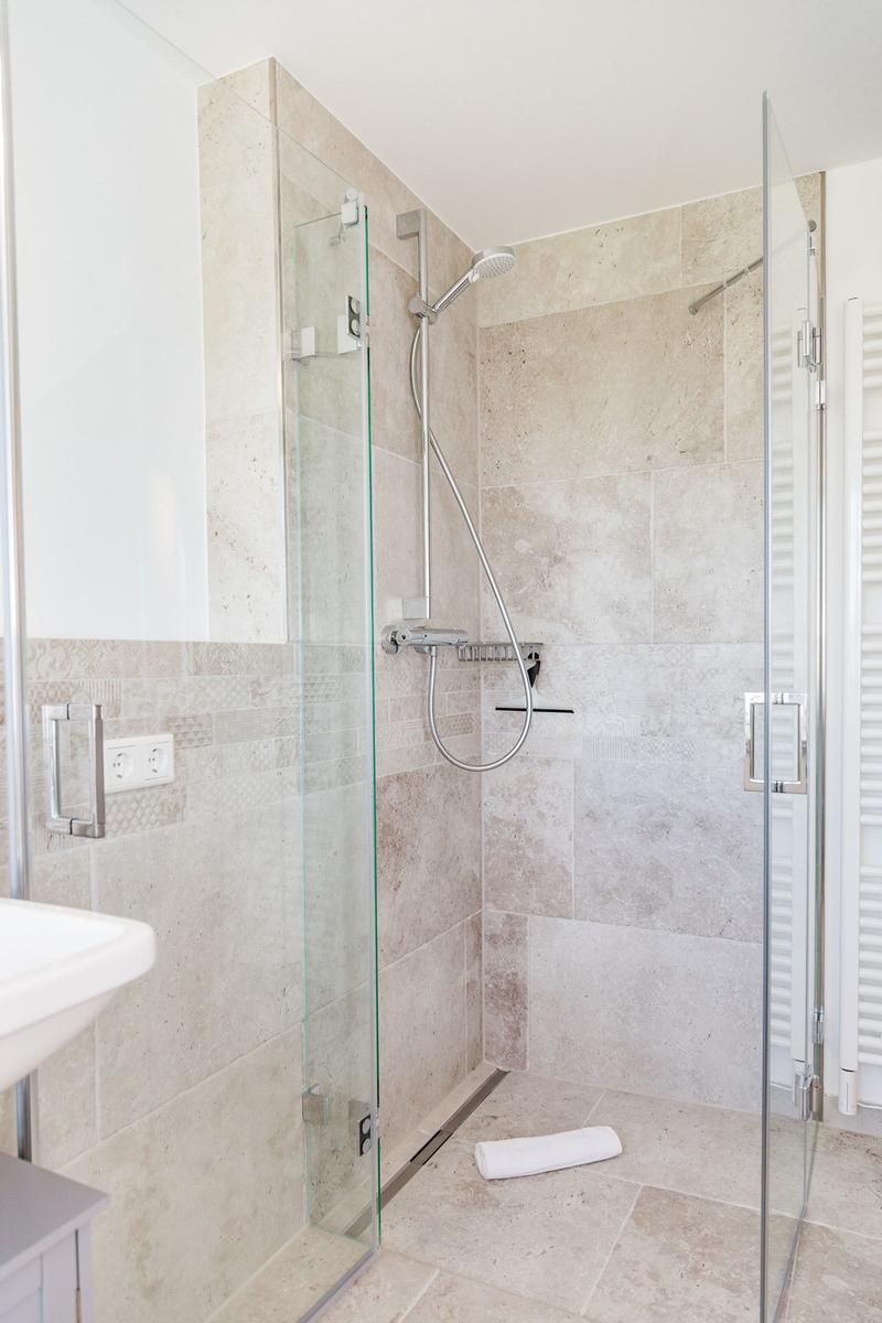 Dusche mit Glaswand, Steinfliesen und Handbrause. Handtuch liegt auf dem Boden.