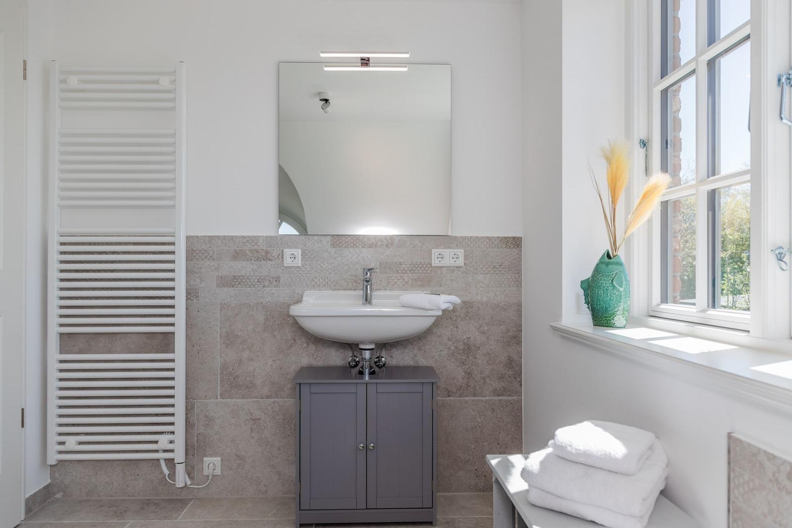 Badezimmer mit Waschbecken, Spiegel, Heizkörper und Fenster mit Vasen.