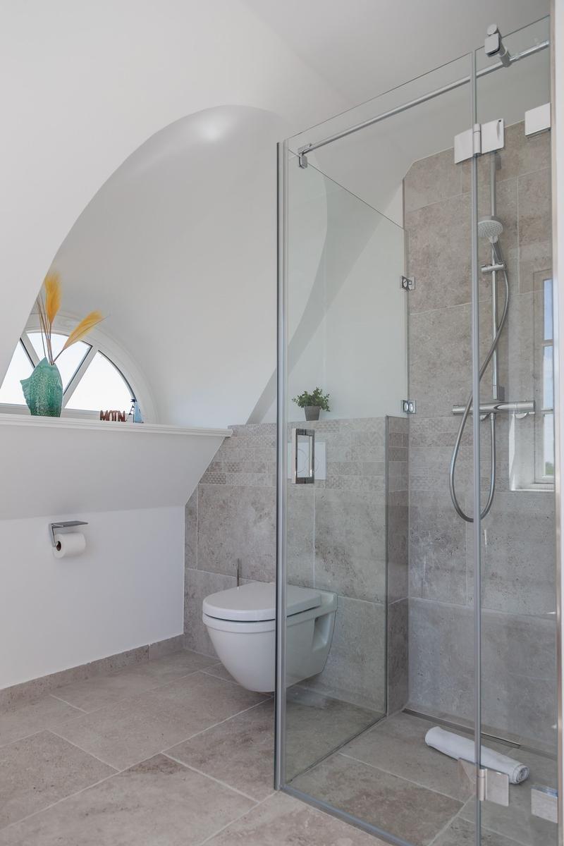 Badezimmer mit Dusche, Toilette und runder Fensteröffnung.