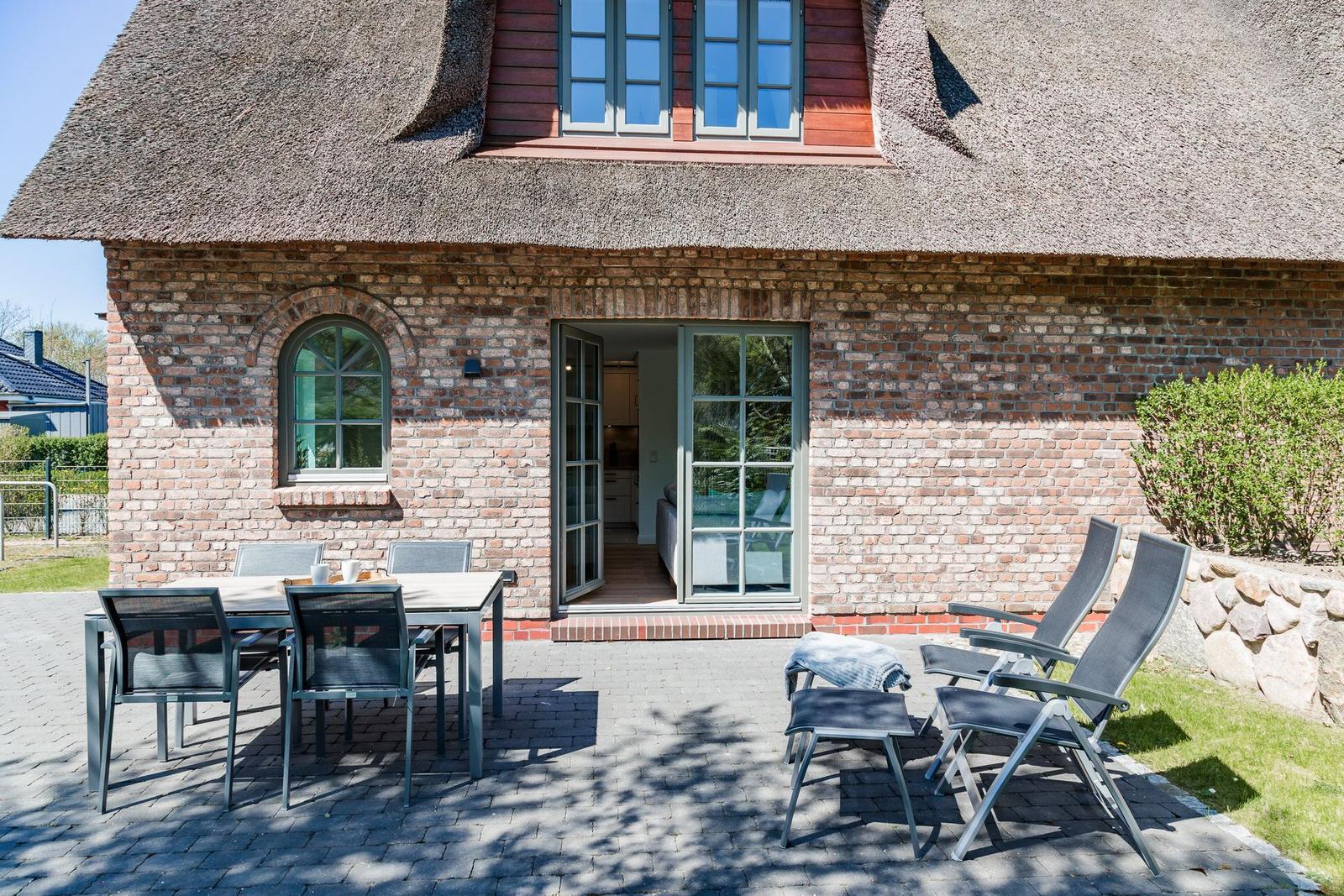 Haus mit Strohdach, Terrasse mit Tisch und Stühlen, Gartentür und Gartenmöbel.