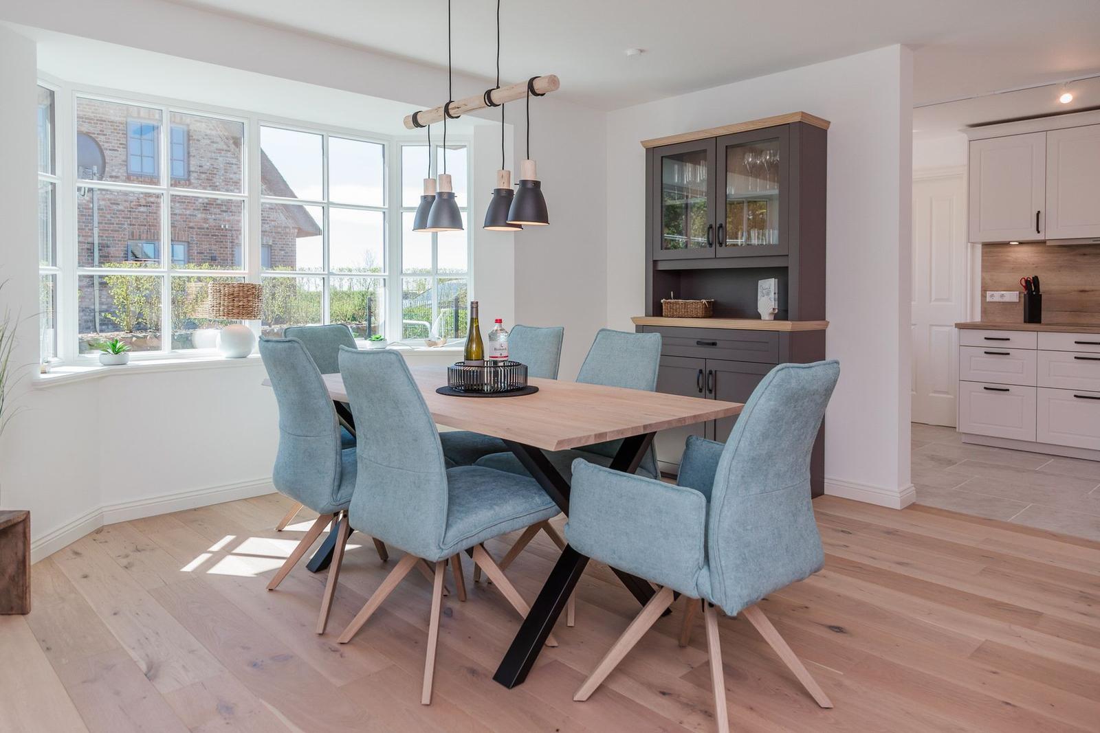 Esszimmer mit Holztisch, sechs blauen Stühlen und großem Fenster. Küchenbereich ist sichtbar.