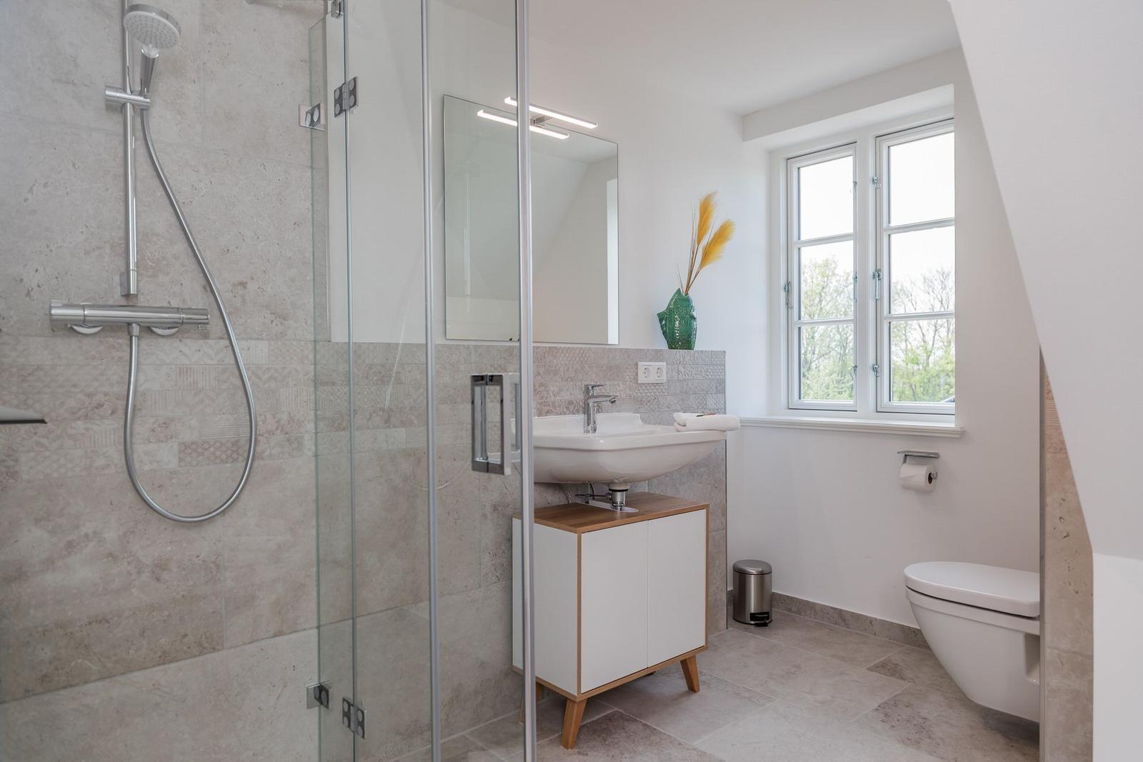 Badezimmer mit Dusche, Waschbecken und Toilette. Fenster mit Blick auf Grün.