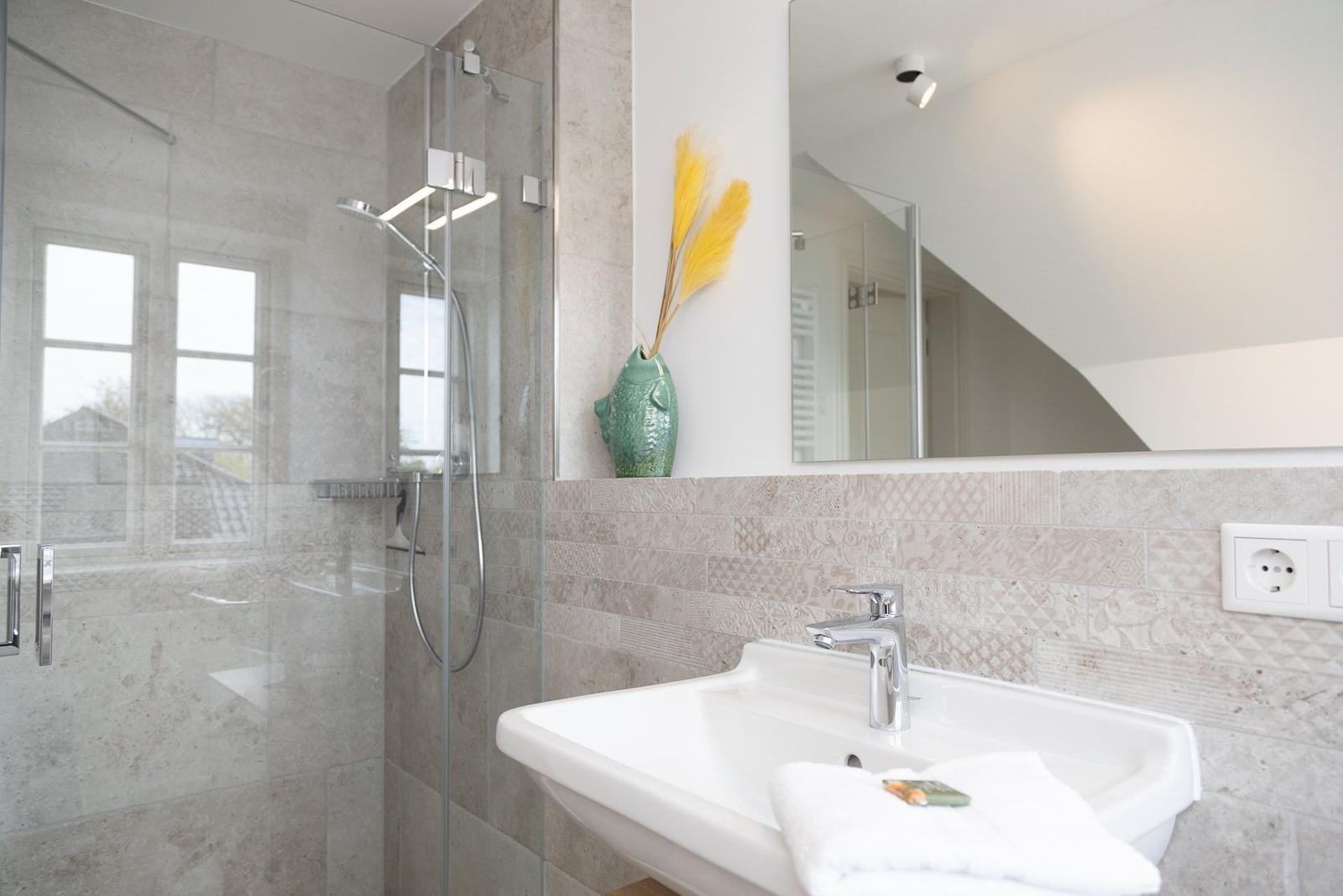 Bad mit Dusche, Waschbecken und Spiegel. Auf Wand hängt grüner Vase mit gelben Pflanzen.