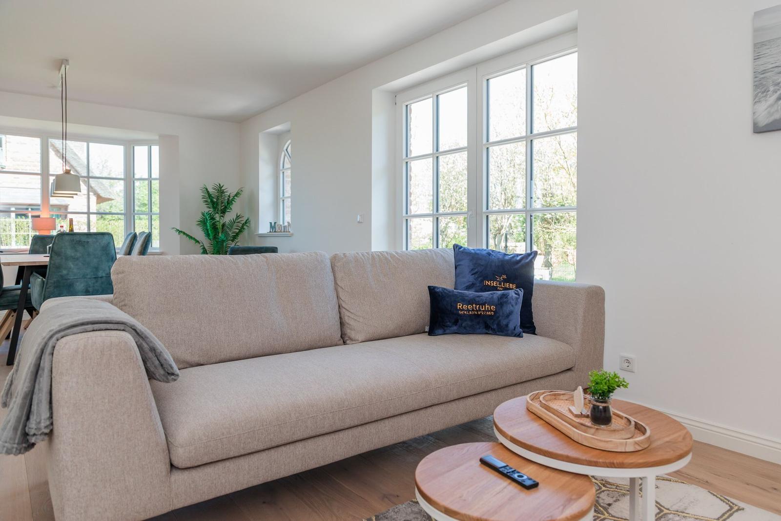 Ein Wohnzimmer mit Sofa, Couchtisch und Fenstern. Auf dem Sofa liegen Kissen.