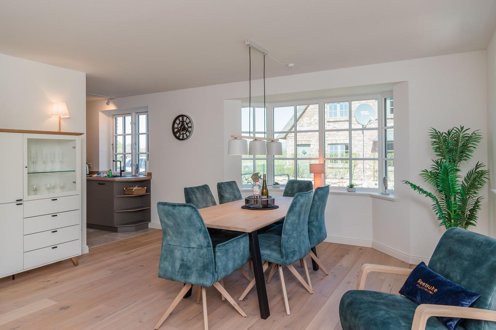Esszimmer mit Holztisch, sechs blauen Stühlen und großem Fenster. Nebenraum mit Küchenzeile und Glasvitrine.